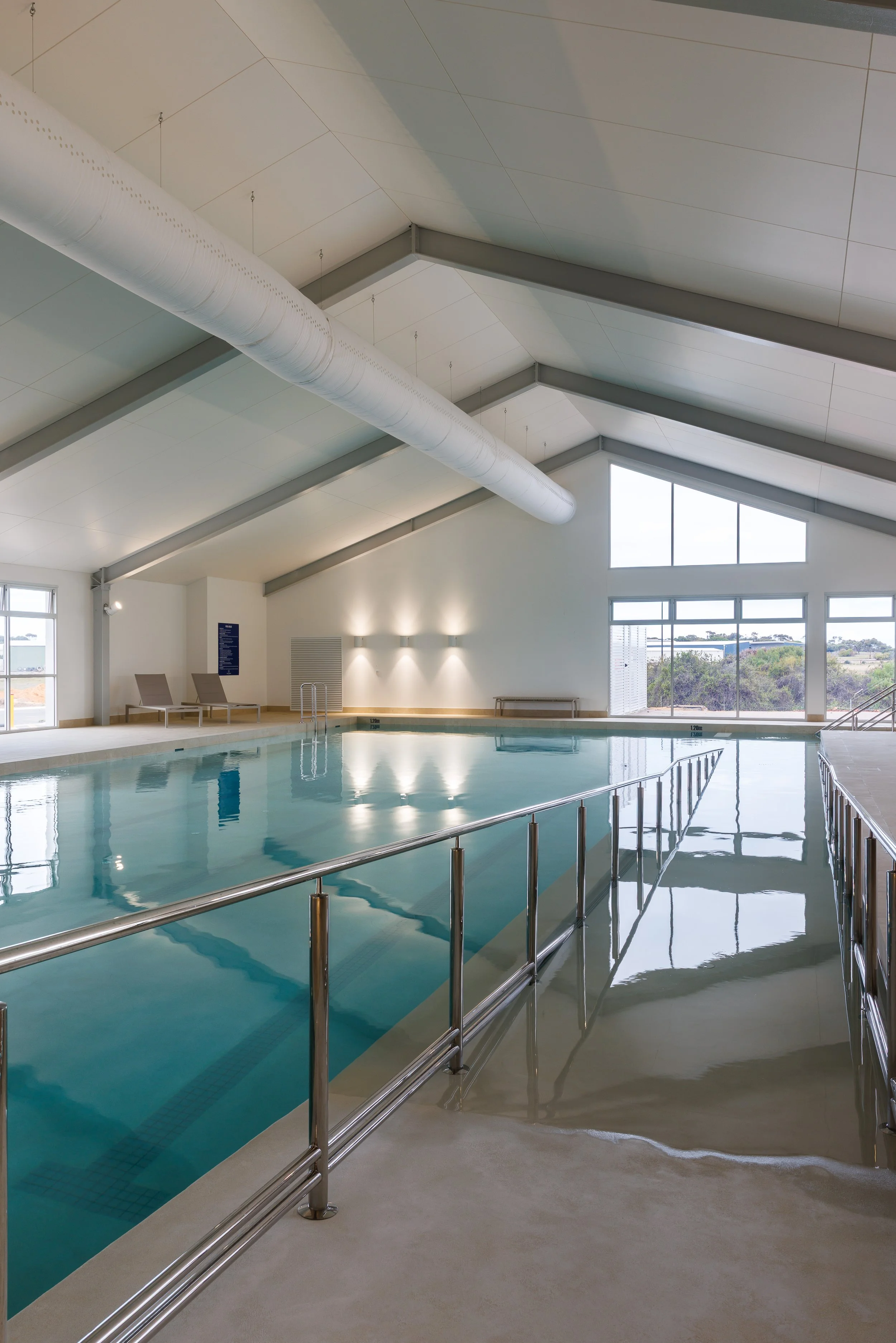 Indoor swimming pool with accessibility handrails, large windows, lounge chairs, and exposed ventilation ducts.