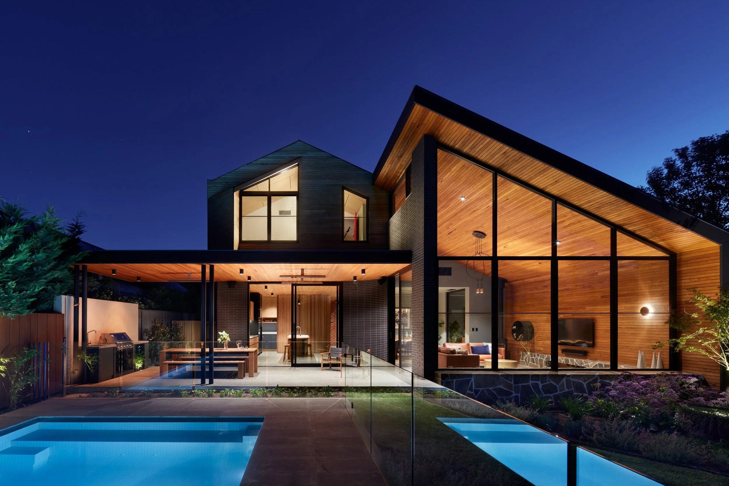 Modern luxury home with wood-paneled vaulted ceilings, floor-to-ceiling windows, outdoor kitchen, and illuminated swimming pool at dusk