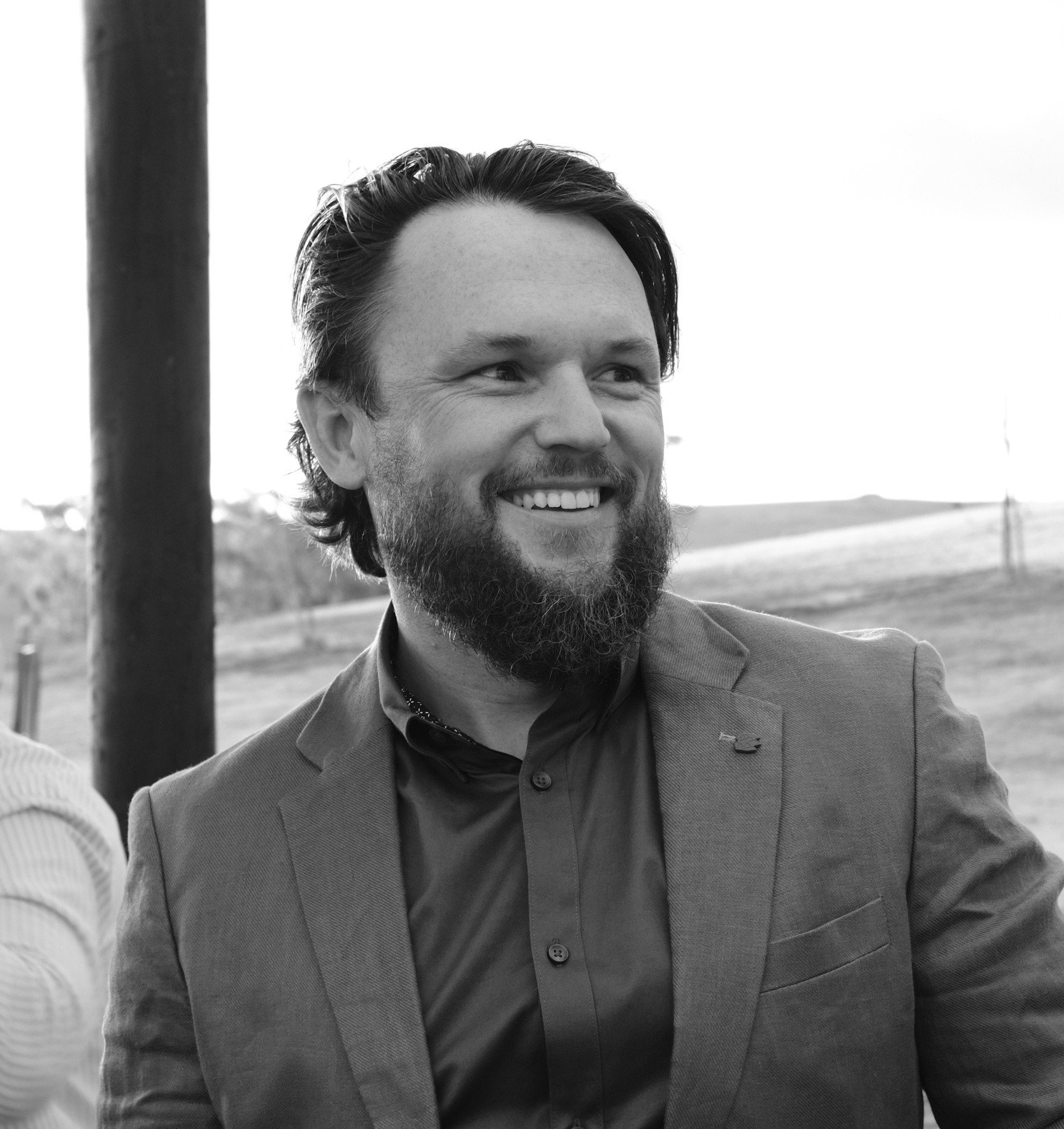 Smiling bearded man in a blazer and button-down shirt, photographed outdoors in black and white