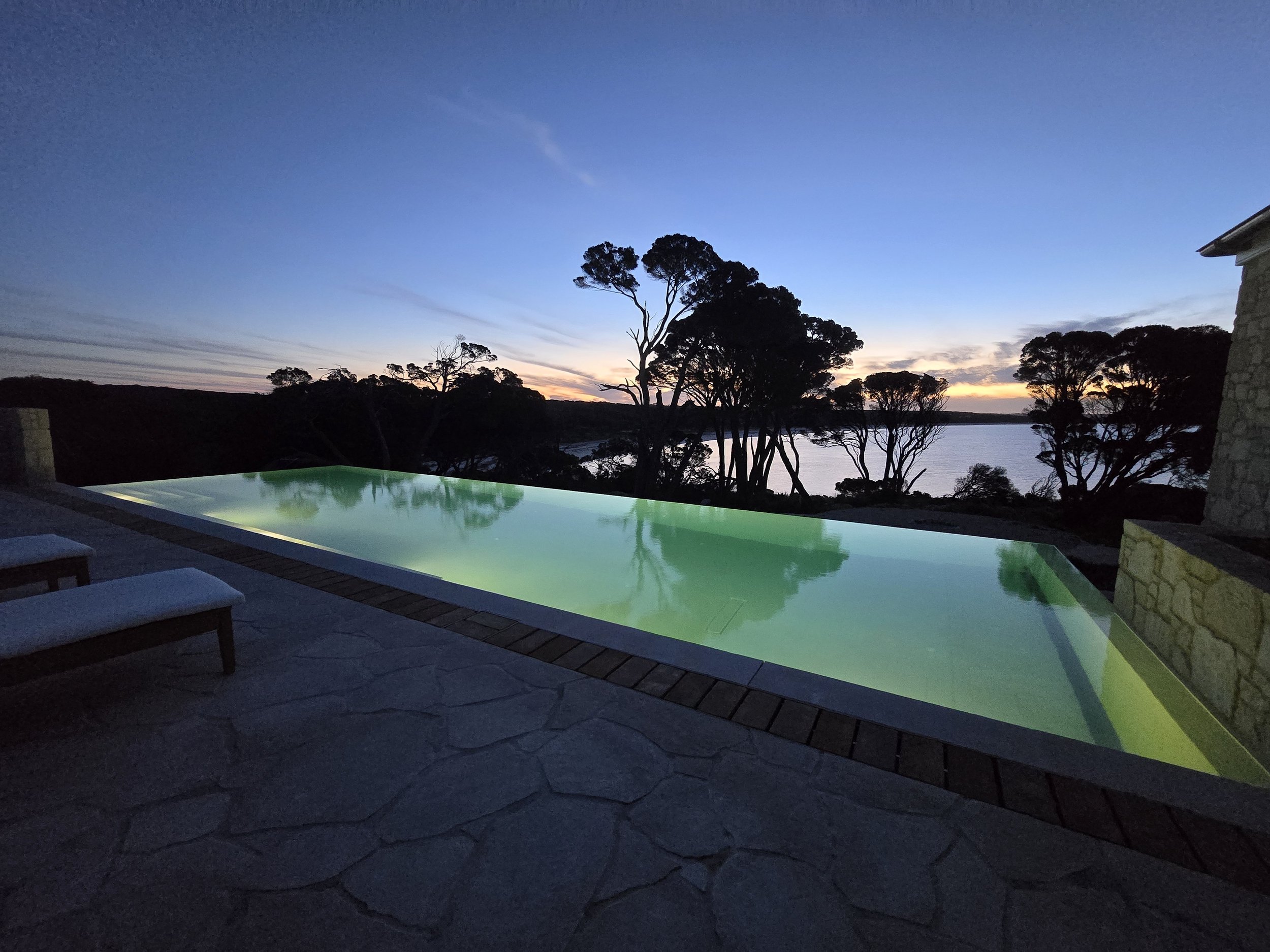 Infinity pool overlooking a calm bay and silhouetted trees at sunset