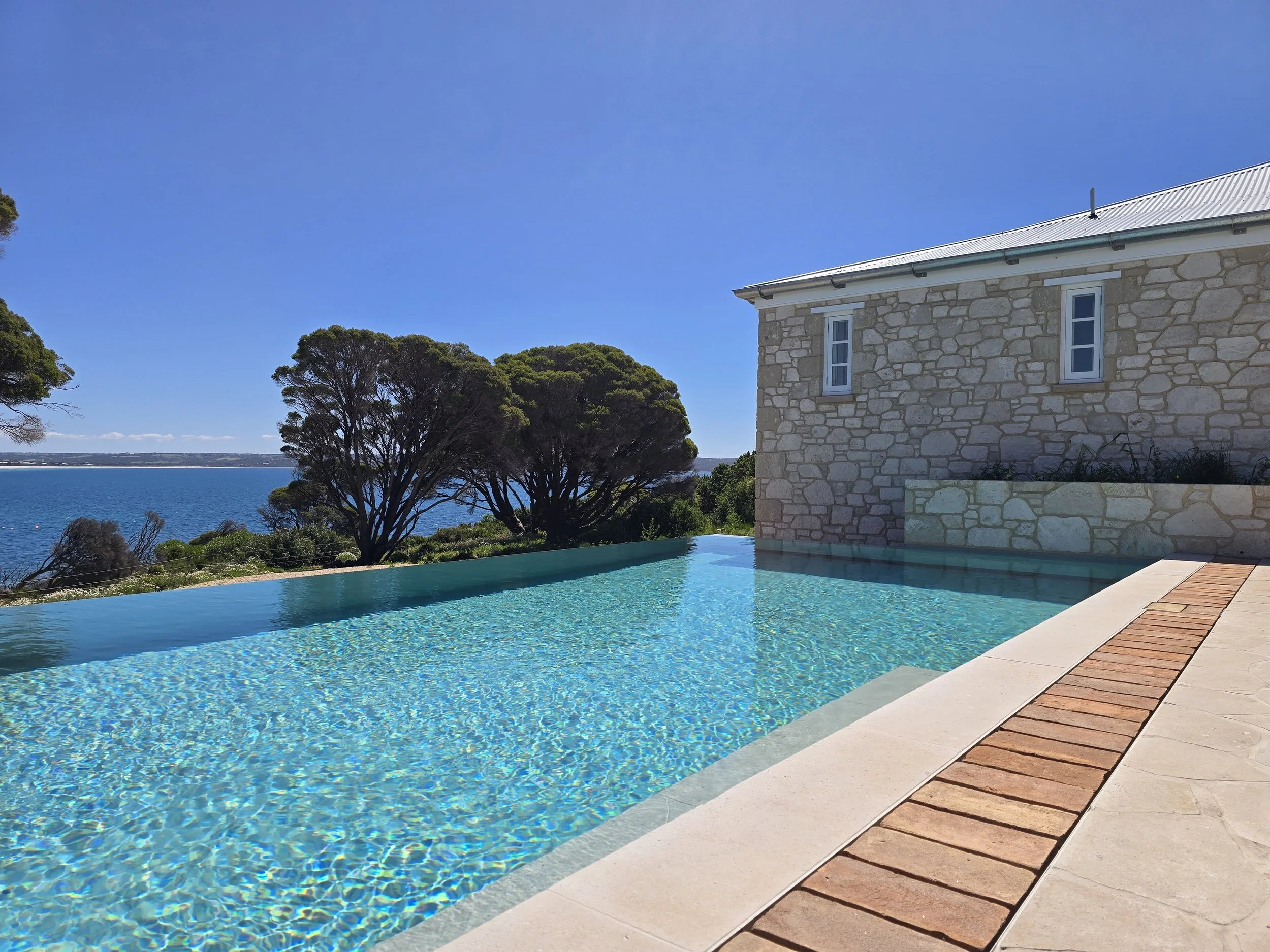 Infinity pool beside a stone house overlooking a blue coastal bay under a clear sky