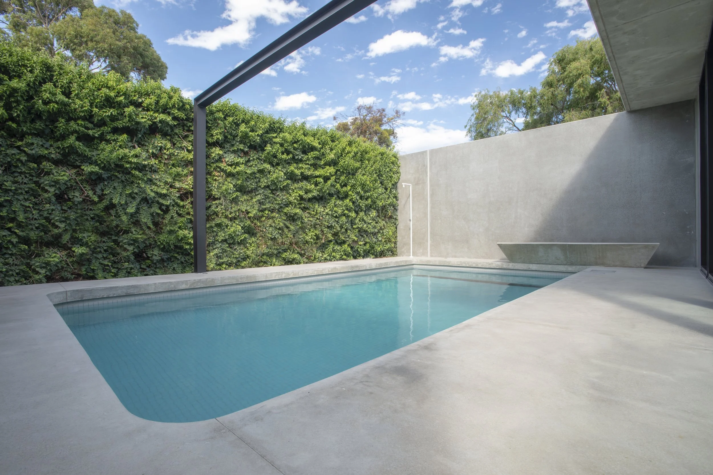 Modern outdoor swimming pool beside a concrete patio, lush greenery, and a minimalist covered terrace under a blue sky