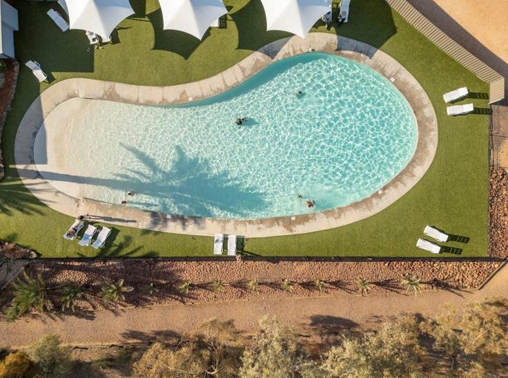 Aerial view of an oval resort swimming pool with swimmers, lounge chairs, umbrellas, and surrounding desert landscaping.