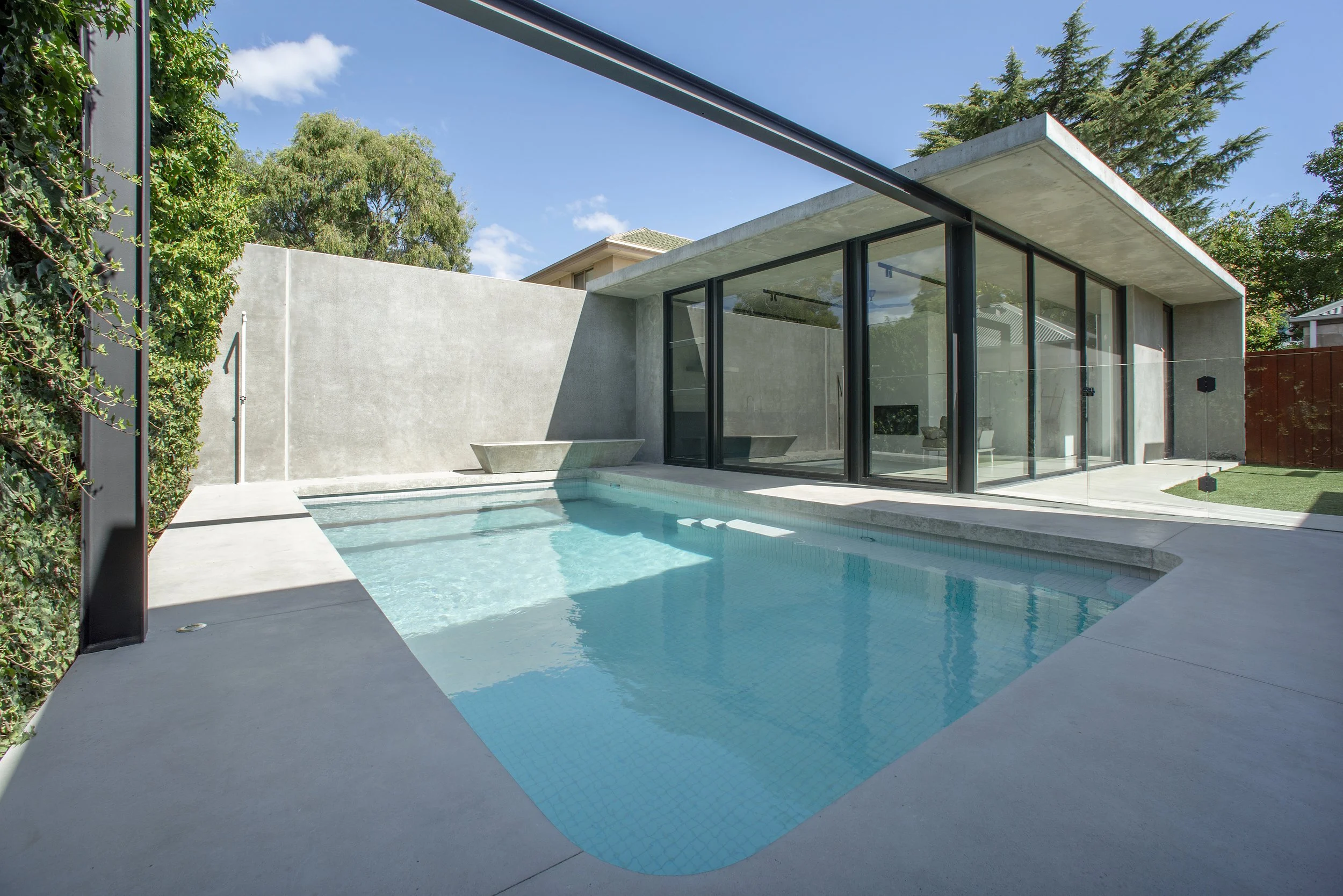 Modern concrete home with floor-to-ceiling windows overlooking a turquoise backyard swimming pool and landscaped patio