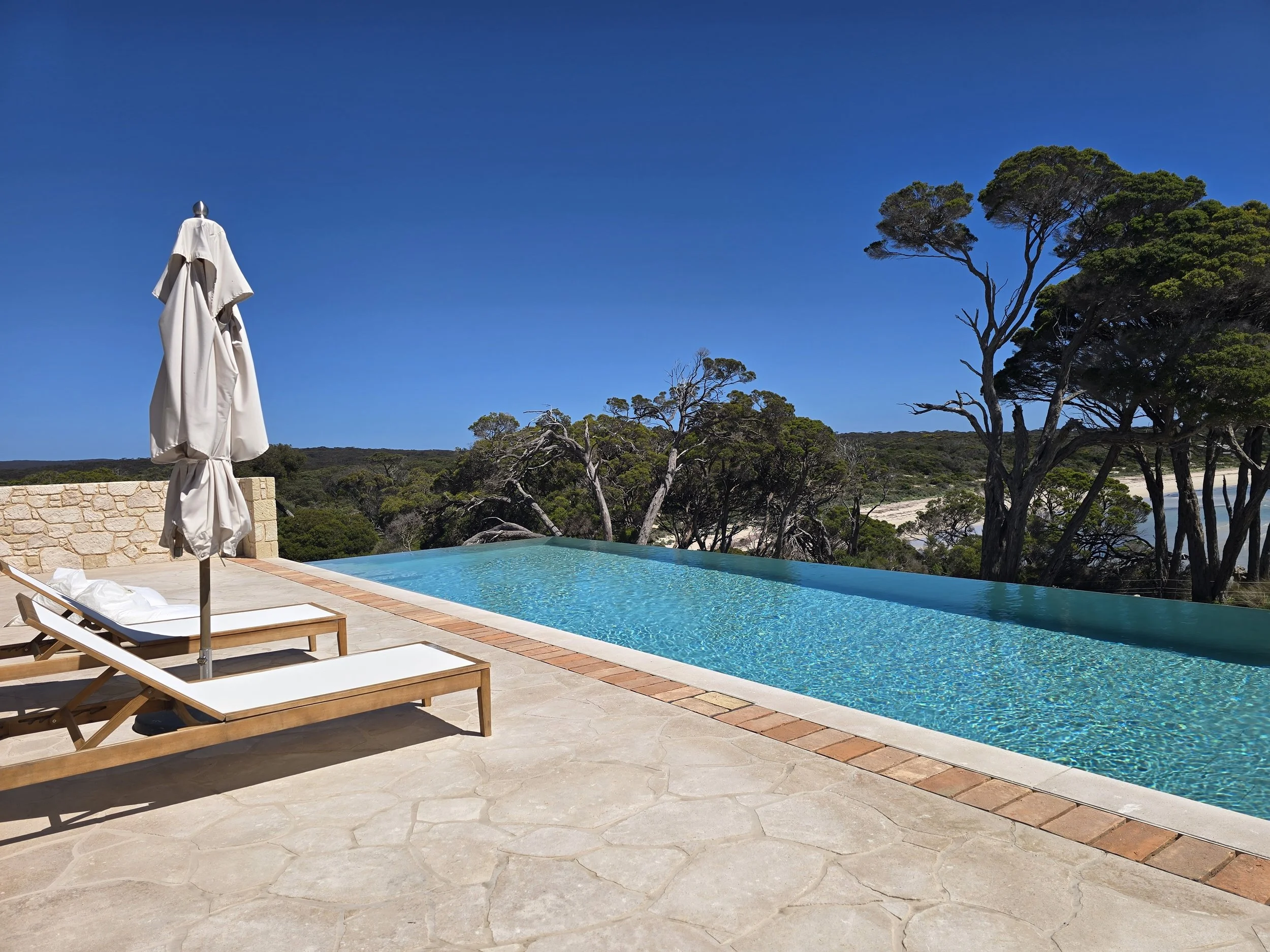Infinity pool overlooking a wooded coastal landscape, with lounge chairs and a closed umbrella on the stone patio.