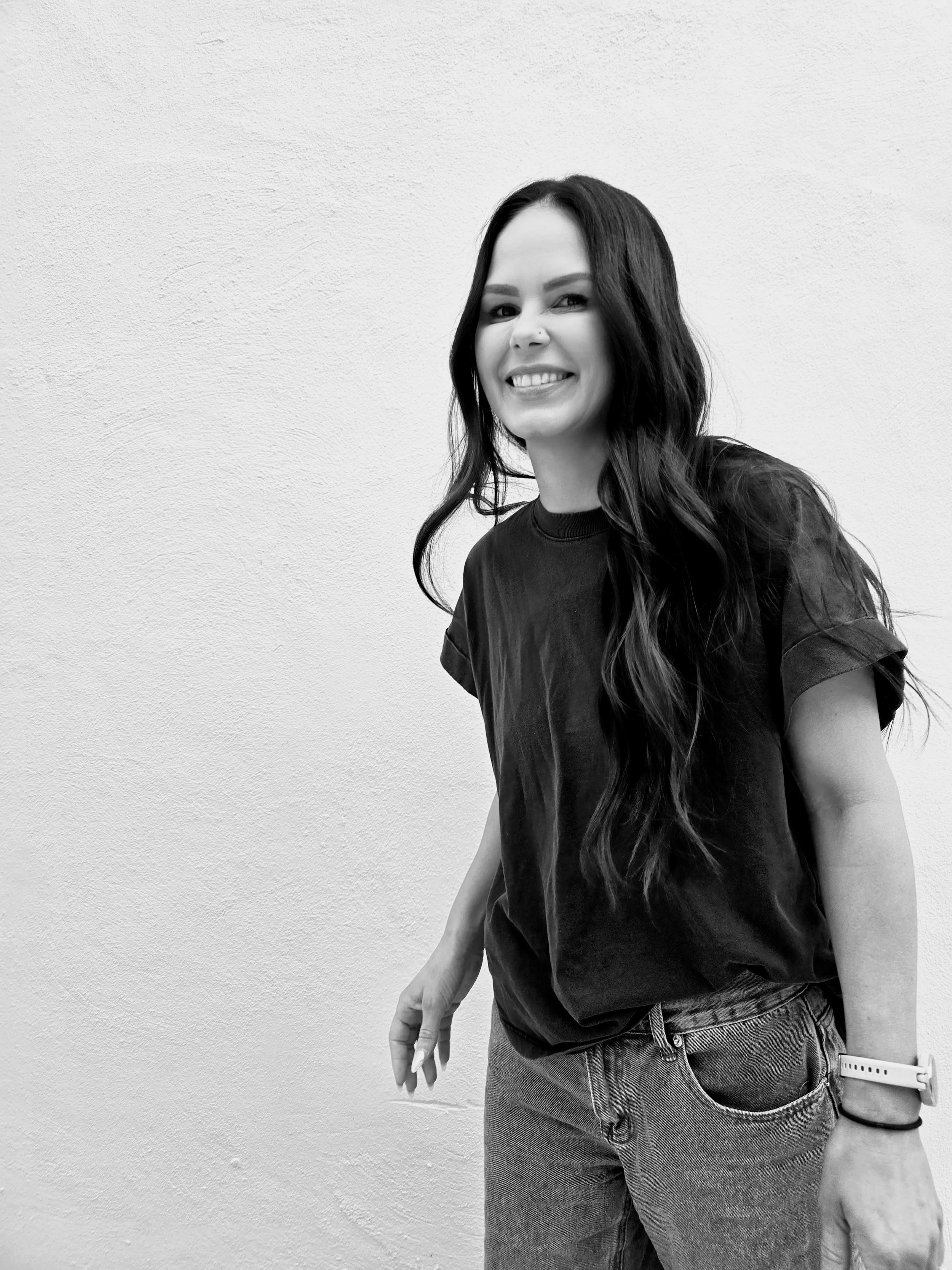 Black-and-white portrait of a smiling woman with long dark hair wearing a T-shirt and jeans against a textured wall.
