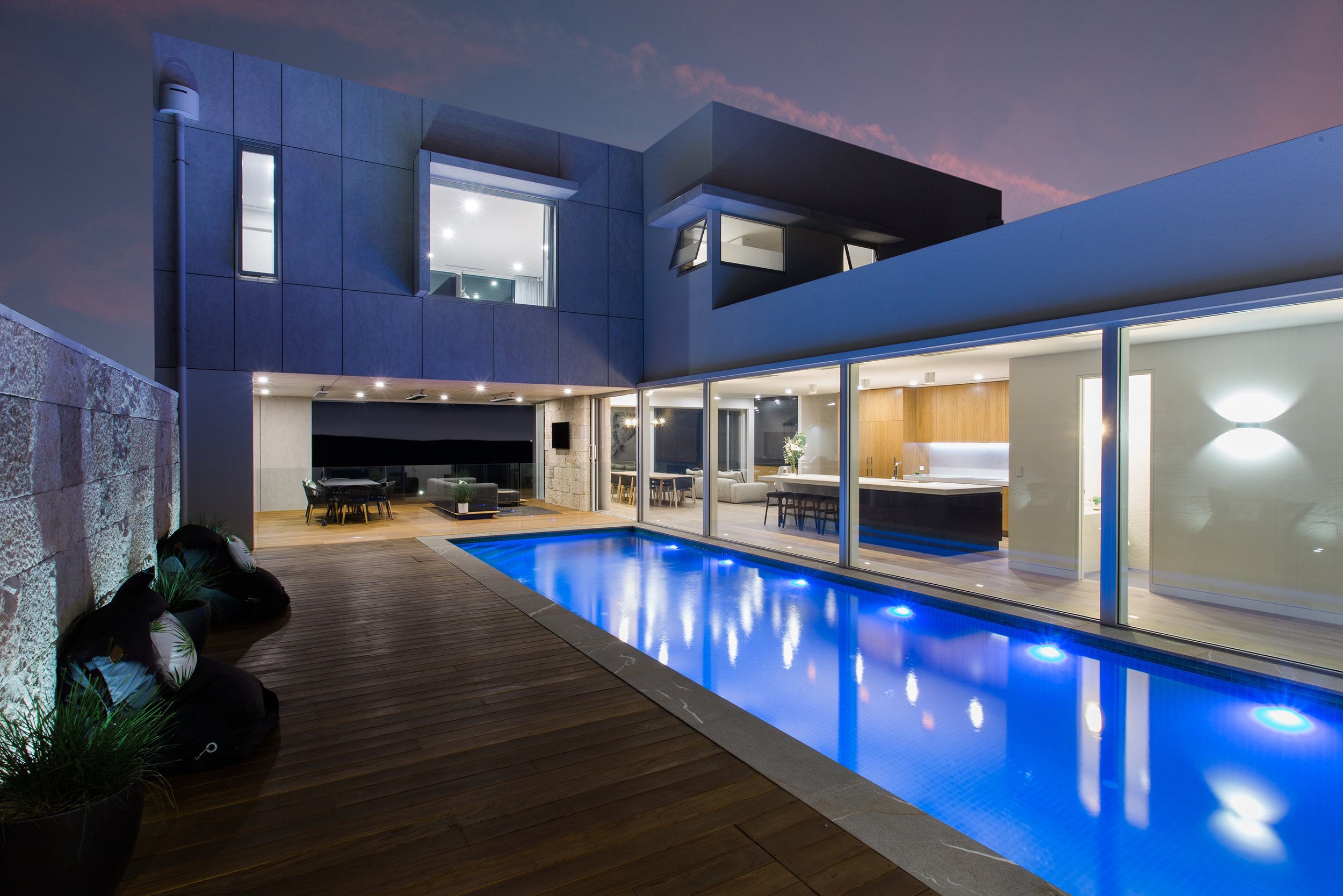 Modern luxury home with illuminated swimming pool, wood deck, floor-to-ceiling windows, and open-plan kitchen at dusk