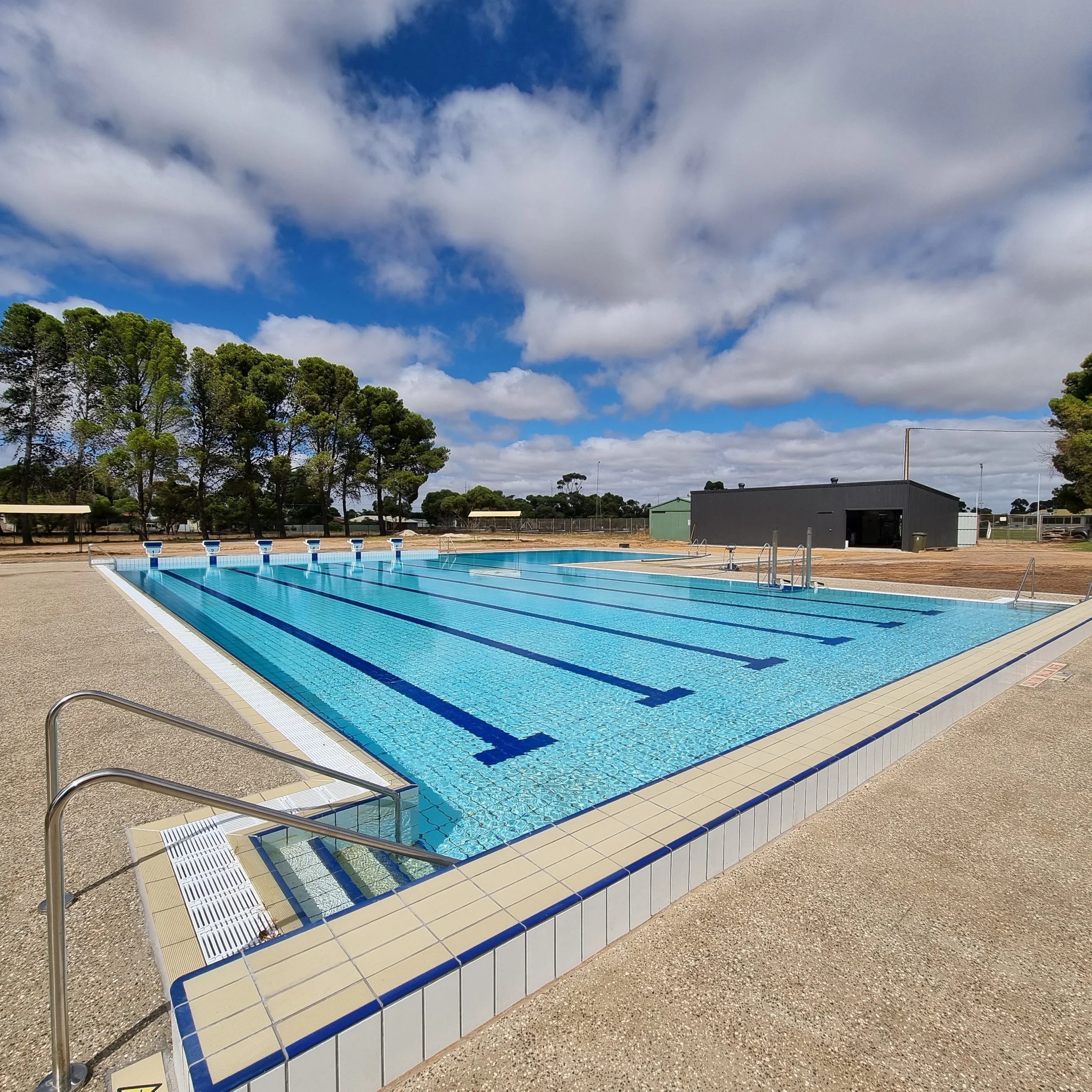 Outdoor lap swimming pool with blue lane markings, starting blocks, ladders, and a cloudy sky