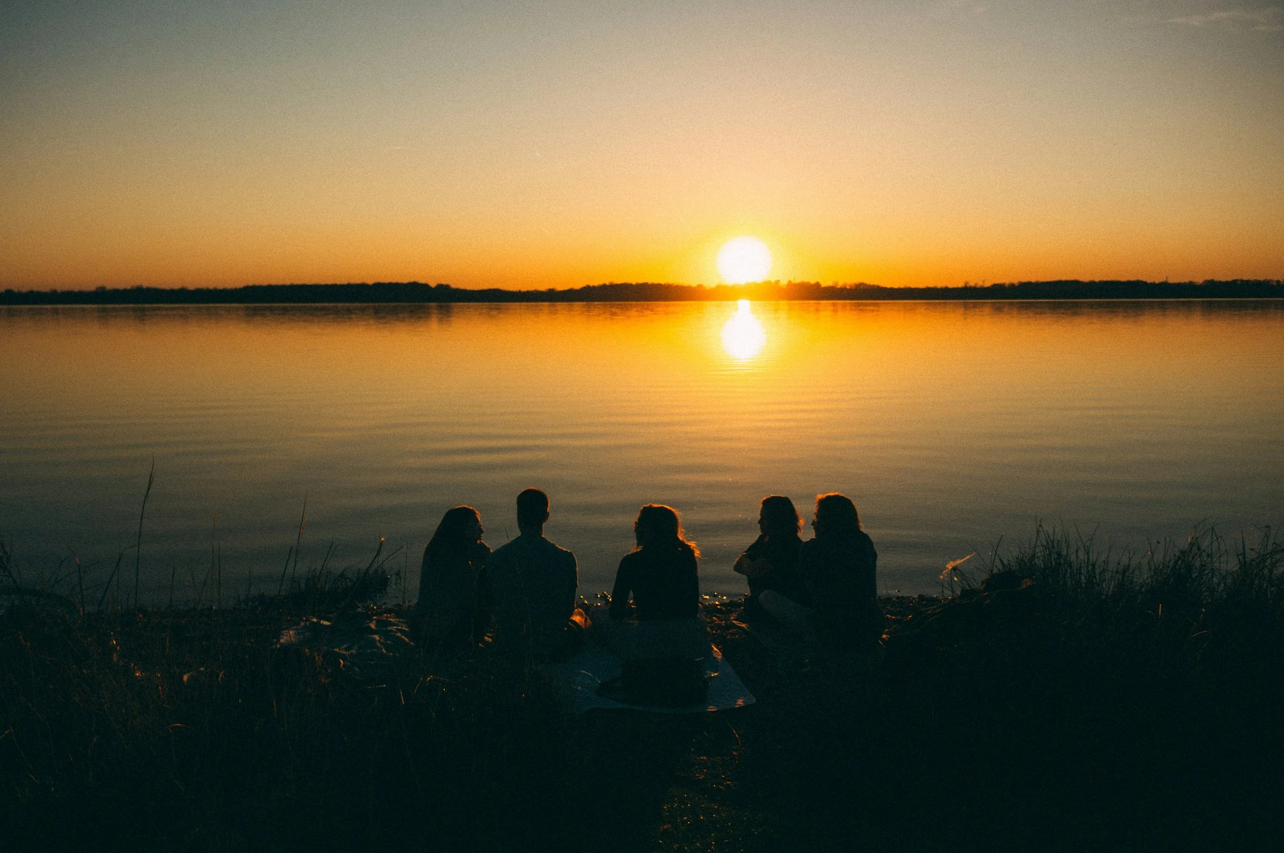 Neljä ihmistä istuvat lähellä veden rantaa auringonlaskun aikaan, taustalla järvi ja horisontti.