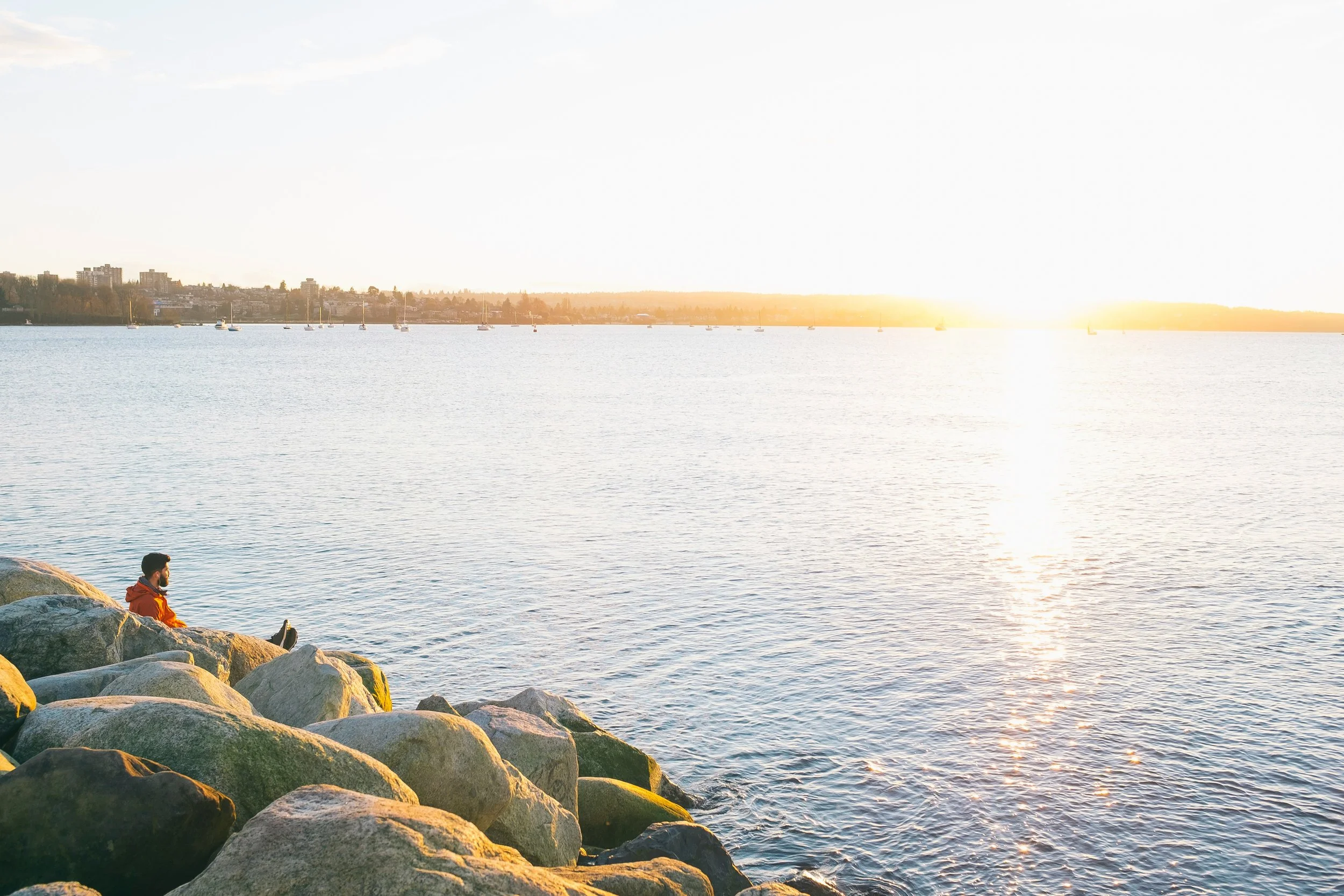 Mies istuu kallioilla katsellen jokea auringonlaskun aikaan, taustalla on veneitä ja kaupunkiä.