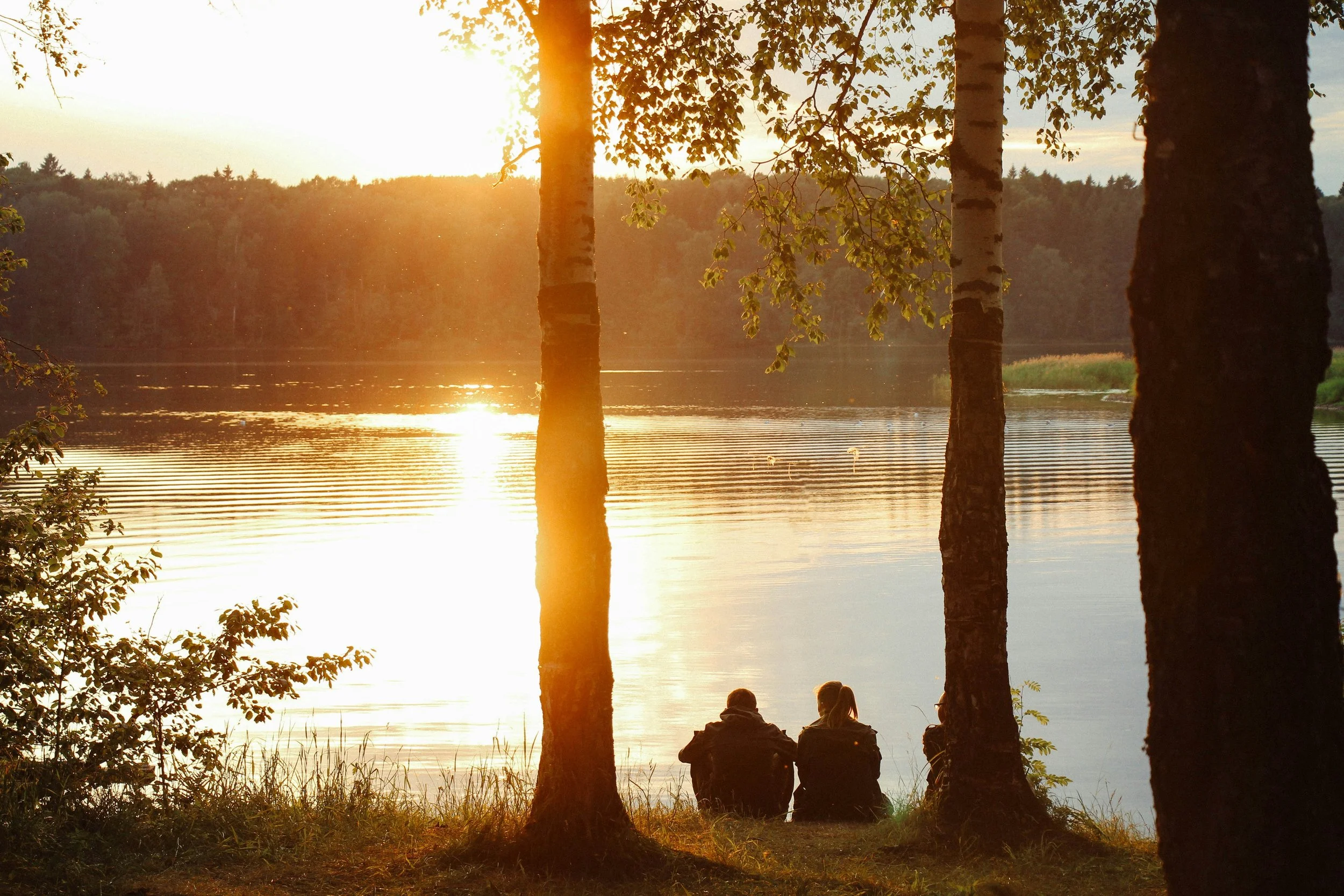 Auringonlasku heijastuu järvestä, kaksi ihmistä istuu rannalla katsomassa näkymää, ympärillä puita ja metsätausta.