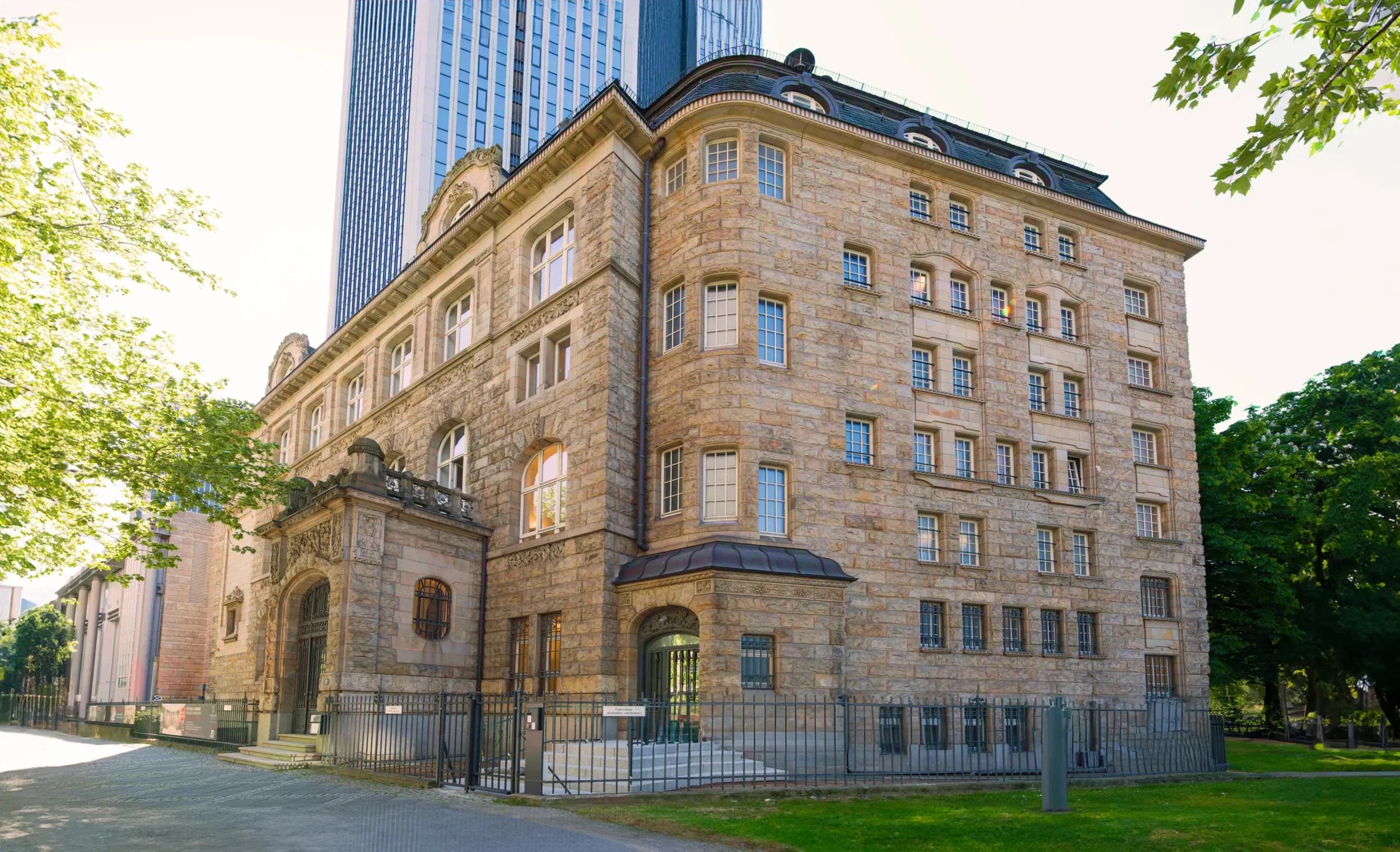 A historic stone building with multiple windows and a rounded corner, surrounded by trees and a low fence in an urban setting.