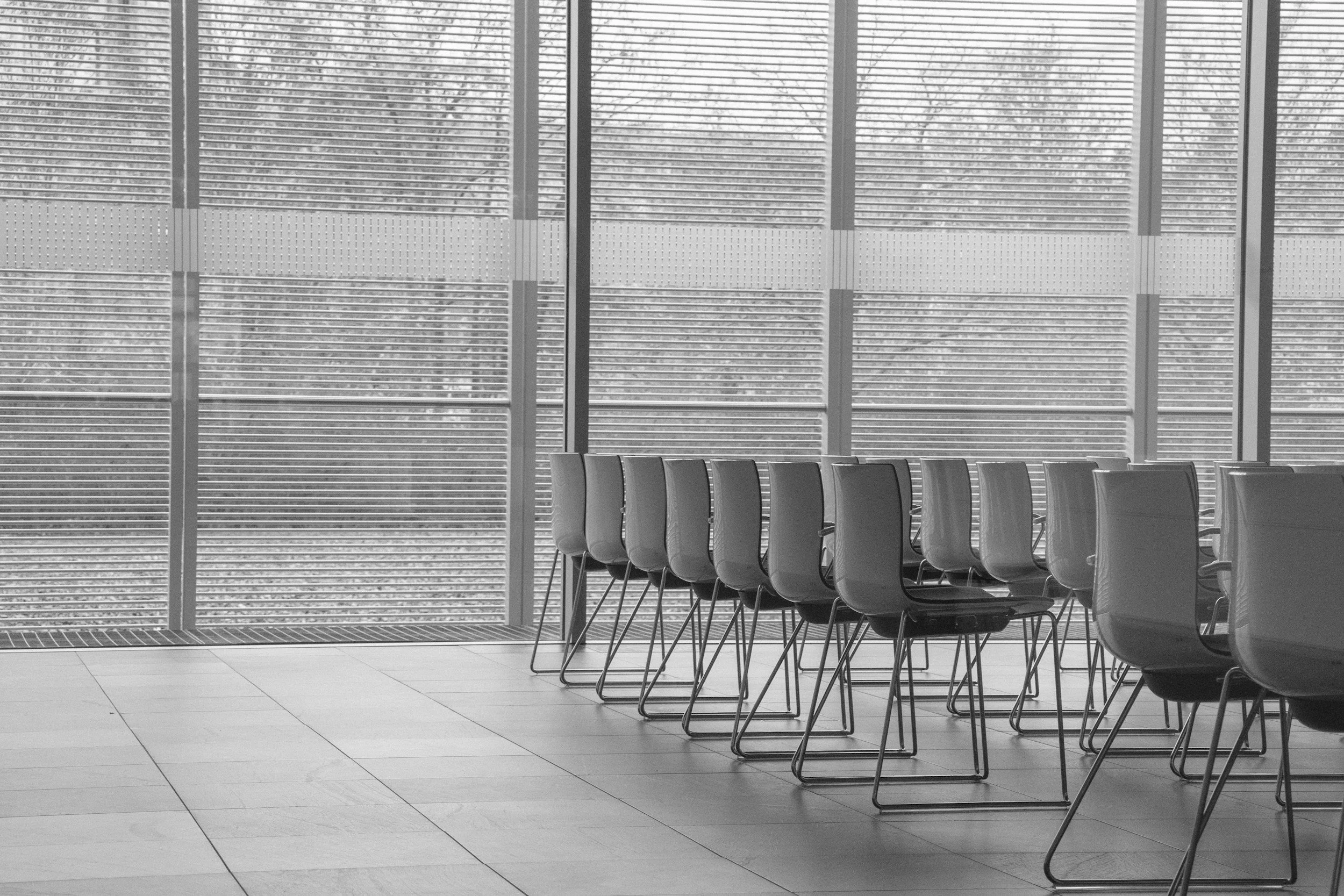 Empty chairs arranged in a row inside a modern building with large glass windows and horizontal blinds.