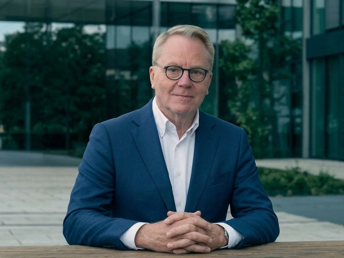 A middle-aged man in a blue suit and white shirt, wearing glasses, is sitting outdoors at a wooden table, with a modern glass building and greenery in the background.