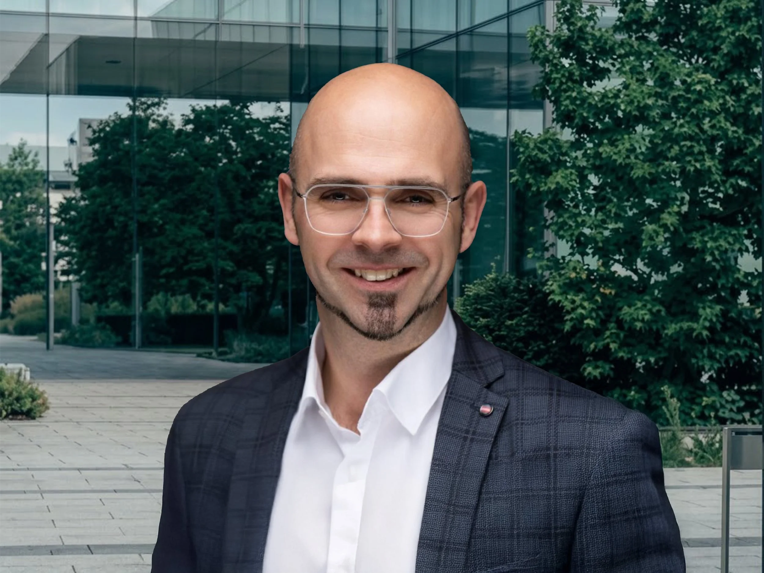 Smiling bald businessman wearing glasses and a dark suit outside a modern glass office building