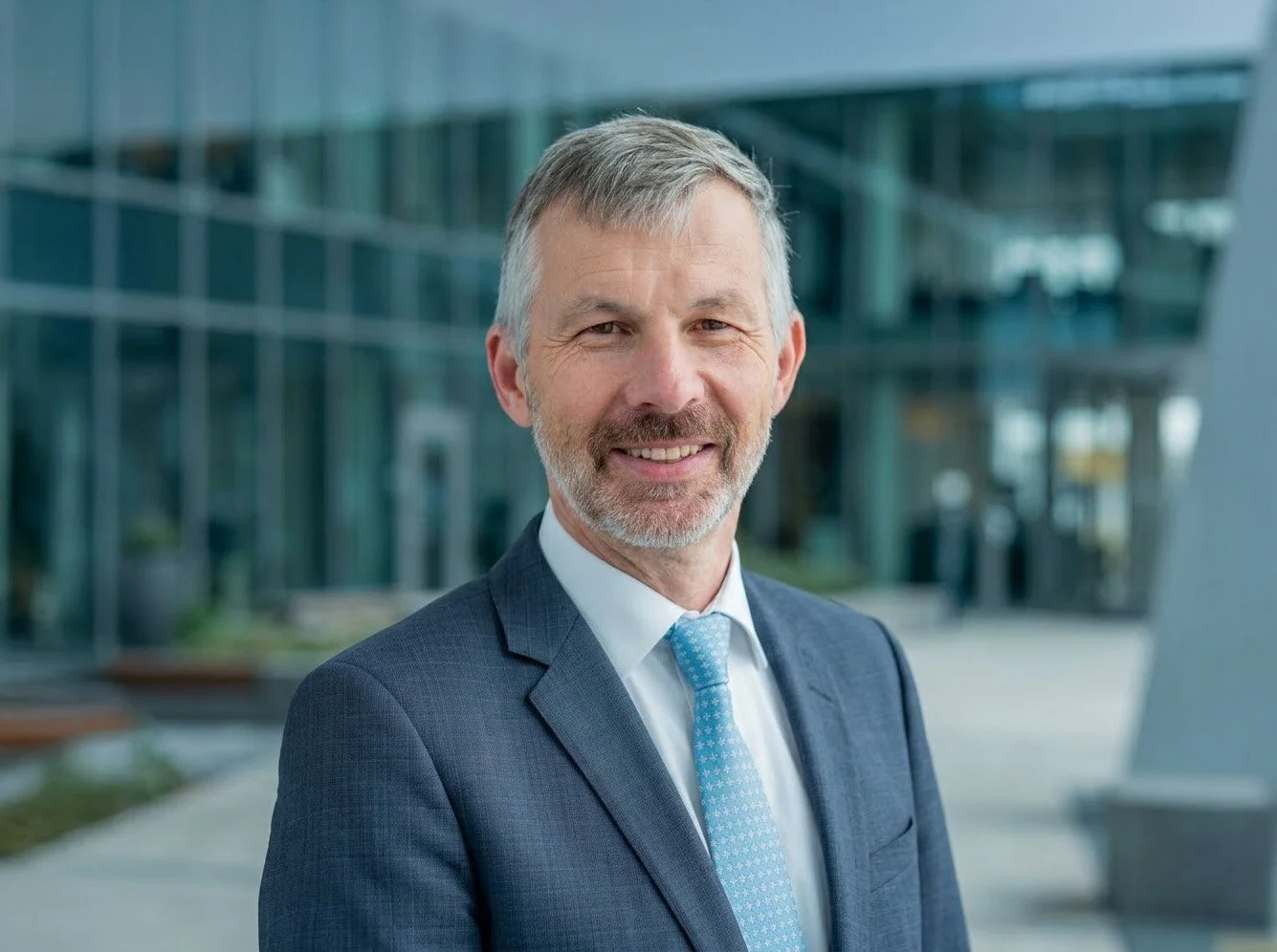 Professional portrait of a middle-aged man in a suit and tie, standing outdoors in front of a modern glass building.