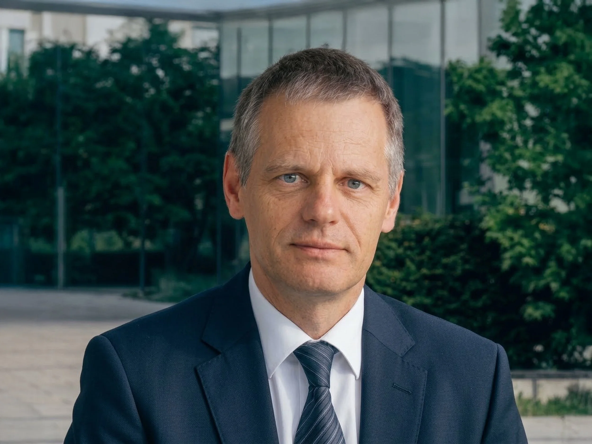 Close-up of a middle-aged man in a suit and tie standing outside in front of a modern glass building with greenery.