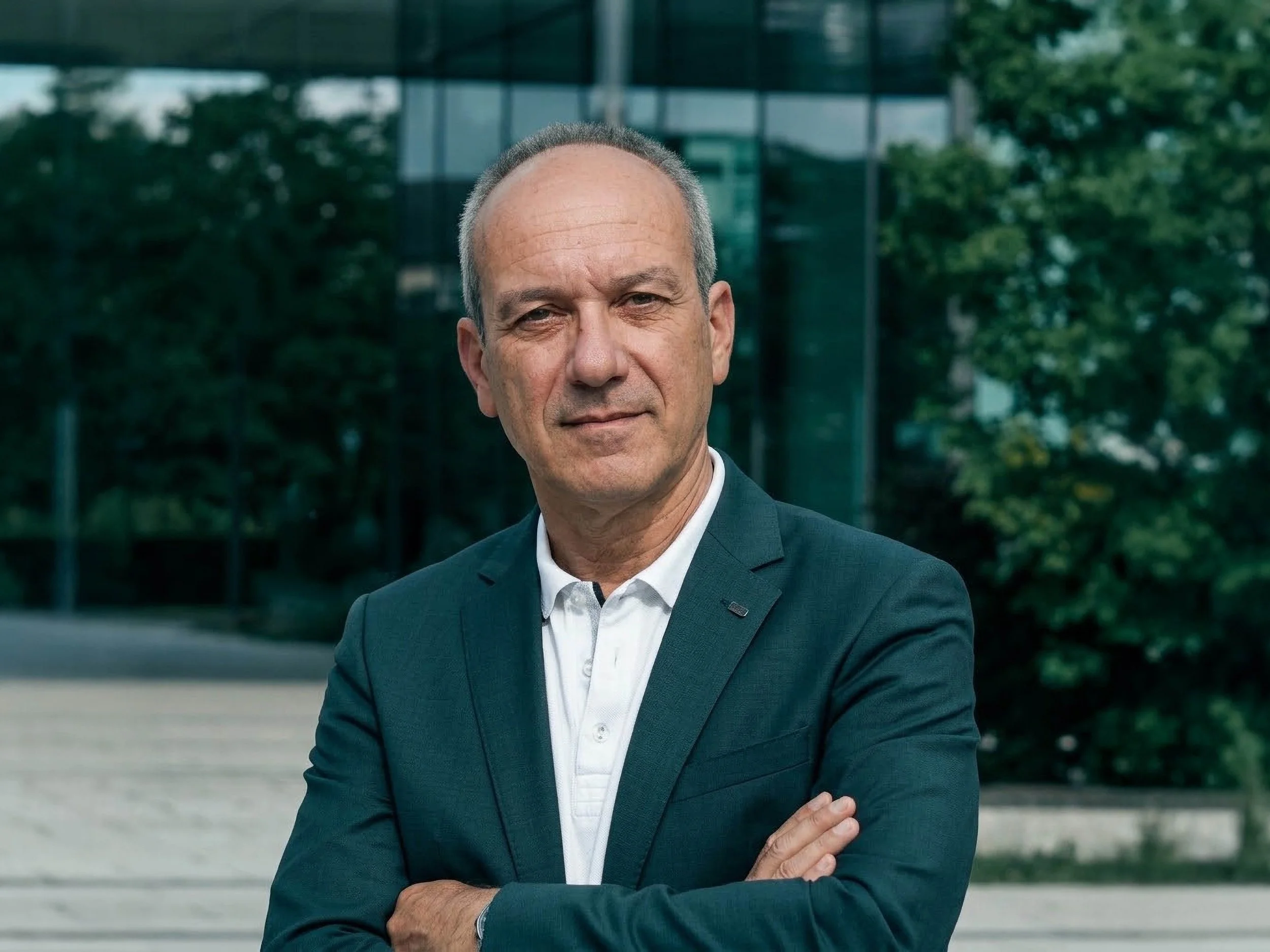 A middle-aged man with short gray hair and a slight smile, wearing a dark suit and white shirt, standing outdoors near modern glass buildings and green trees.
