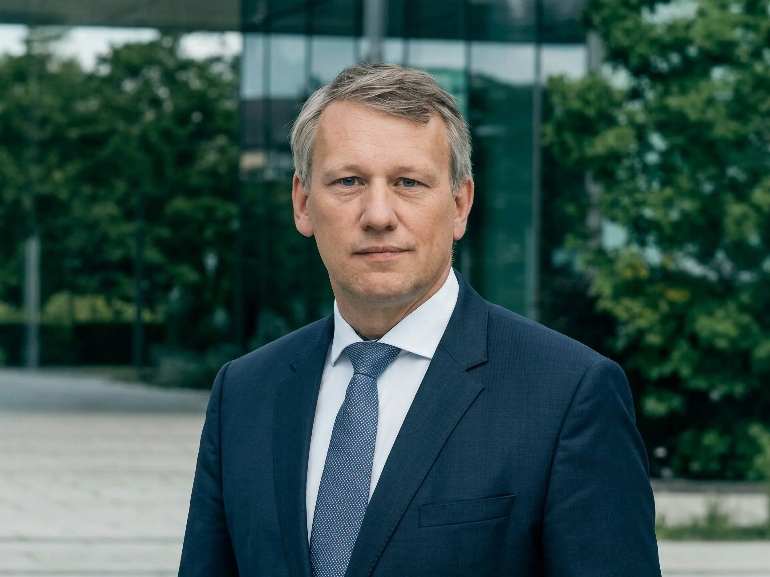 A man in a dark blue suit, white shirt, and patterned tie standing outdoors with modern glass buildings and green trees in the background.