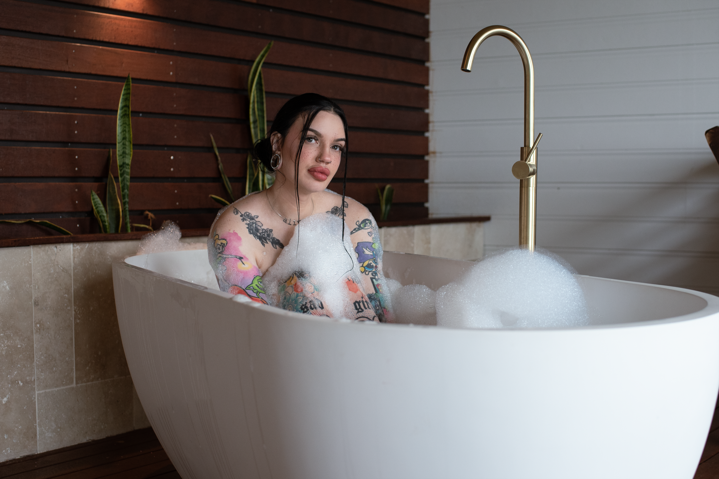 A young woman with dark hair, tattoos, and piercings soaking in a bubble-filled white bathtub in a modern bathroom with a wood-paneled wall and plants.