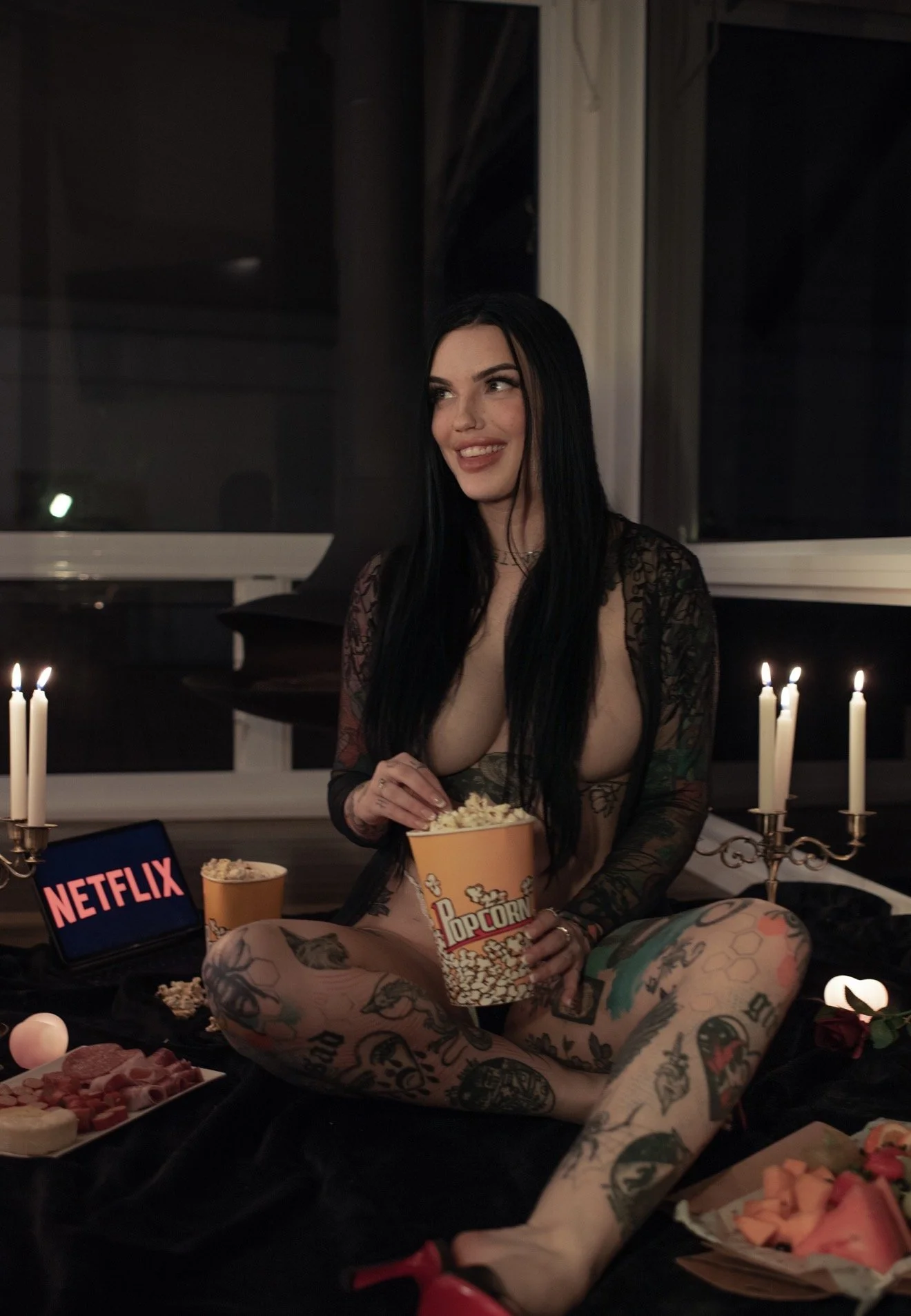 A woman with long black hair and tattoos on her legs and arms, sitting on the floor in a dimly lit room with candles, watching Netflix on a tablet, surrounded by snacks including popcorn and assorted foods.