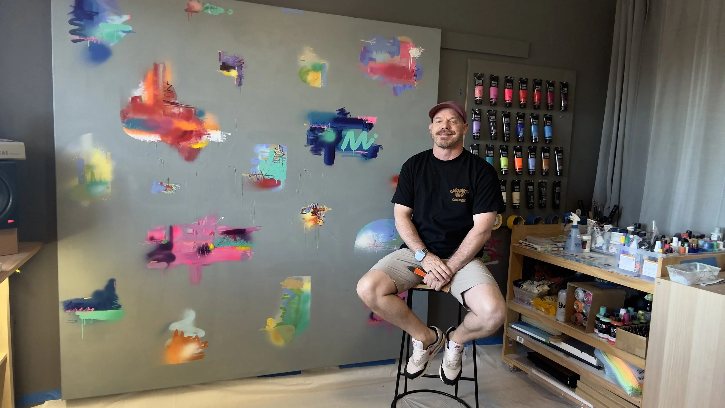 Artist in studio in Sydney on a stool in front of an abstract colorful painting on a large canvas that he has painted