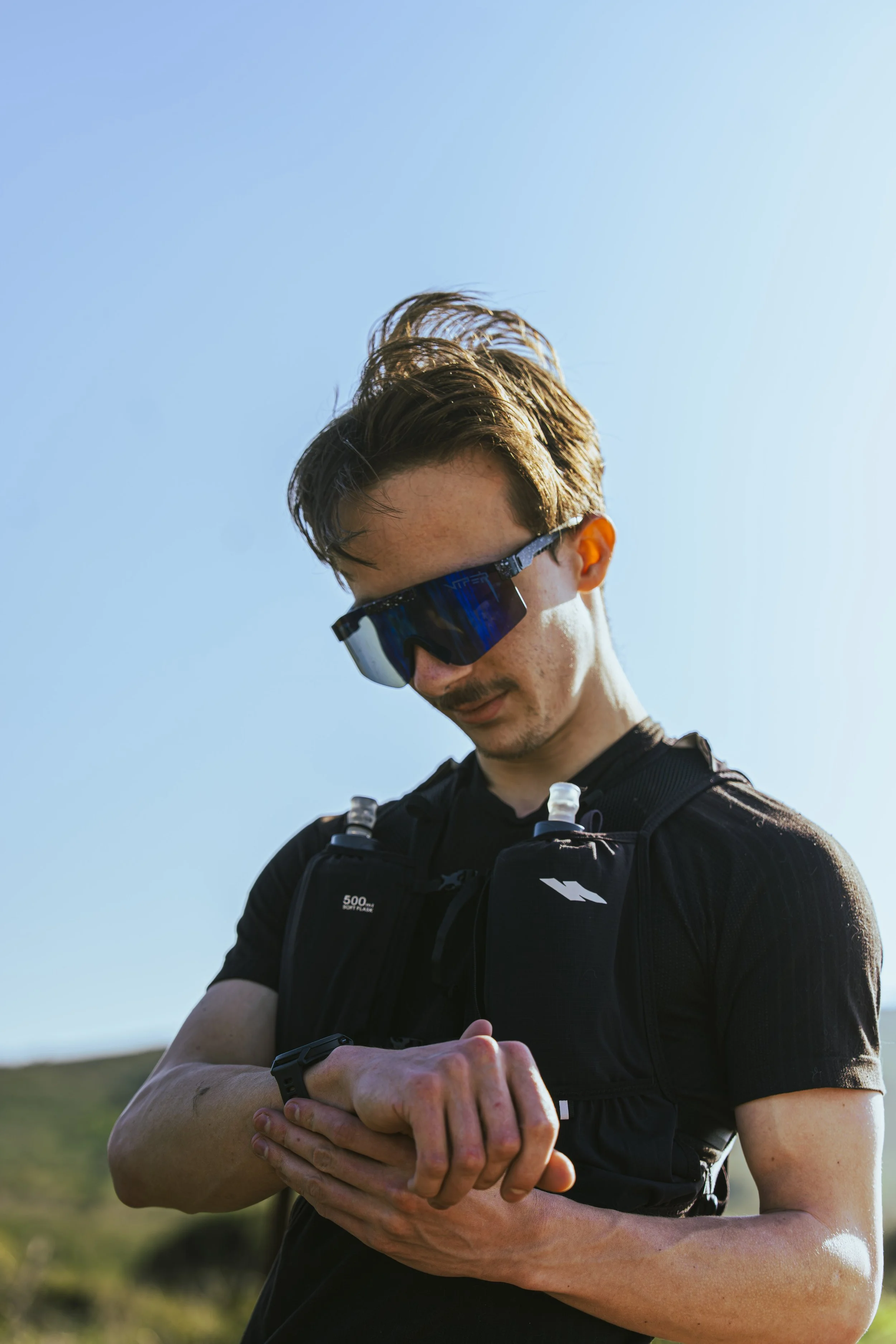 A man with short brown hair and sunglasses, wearing a black athletic shirt and a running vest, is outdoors with a clear blue sky in the background. He is looking down and adjusting his watch or fitness device.