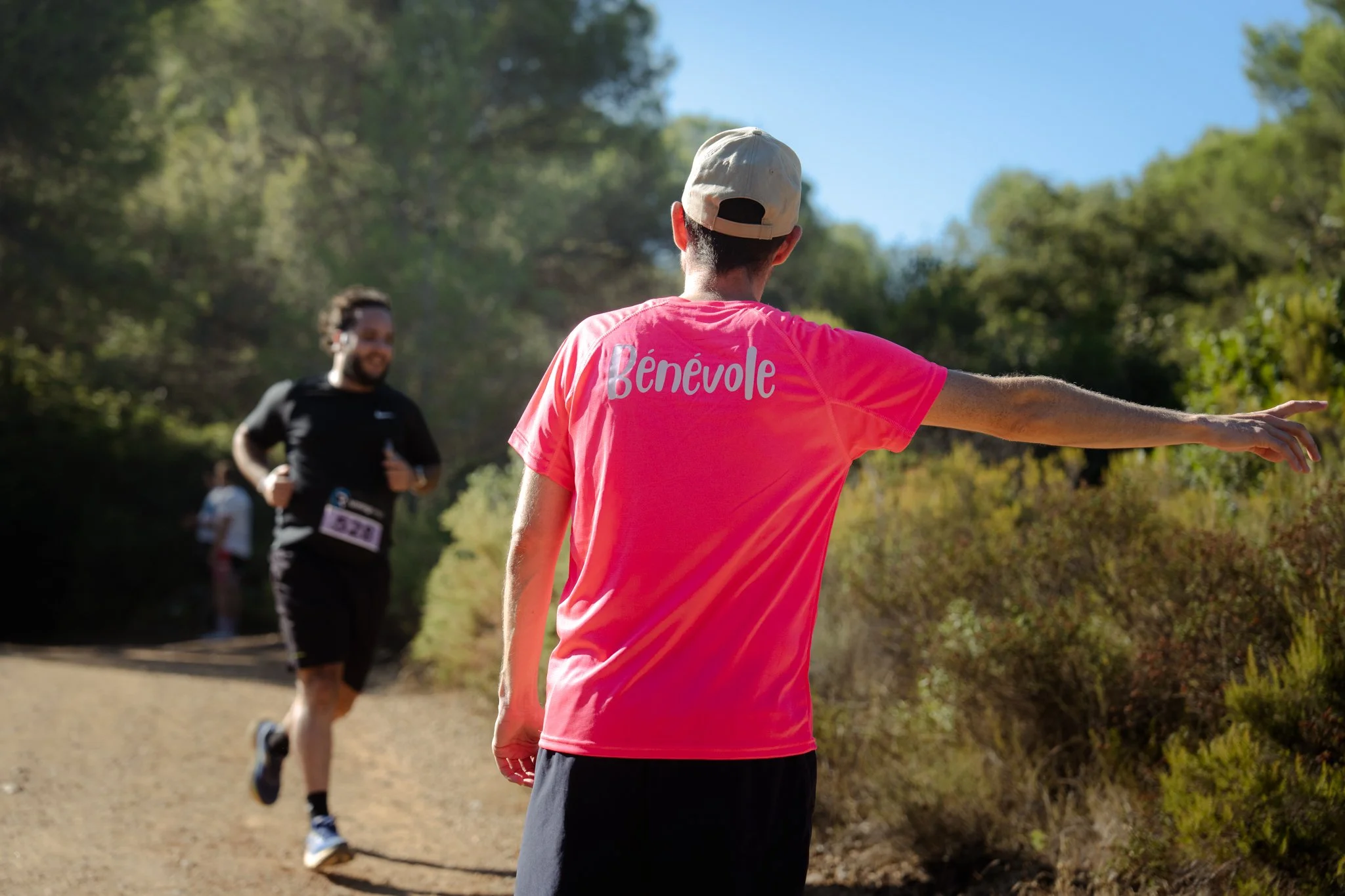 Groupe de personnes courant en plein air sur un sentier en forêt, l'une d'entre elles portant un t-shirt rose avec le mot 'Bénévole' écrit dessus.