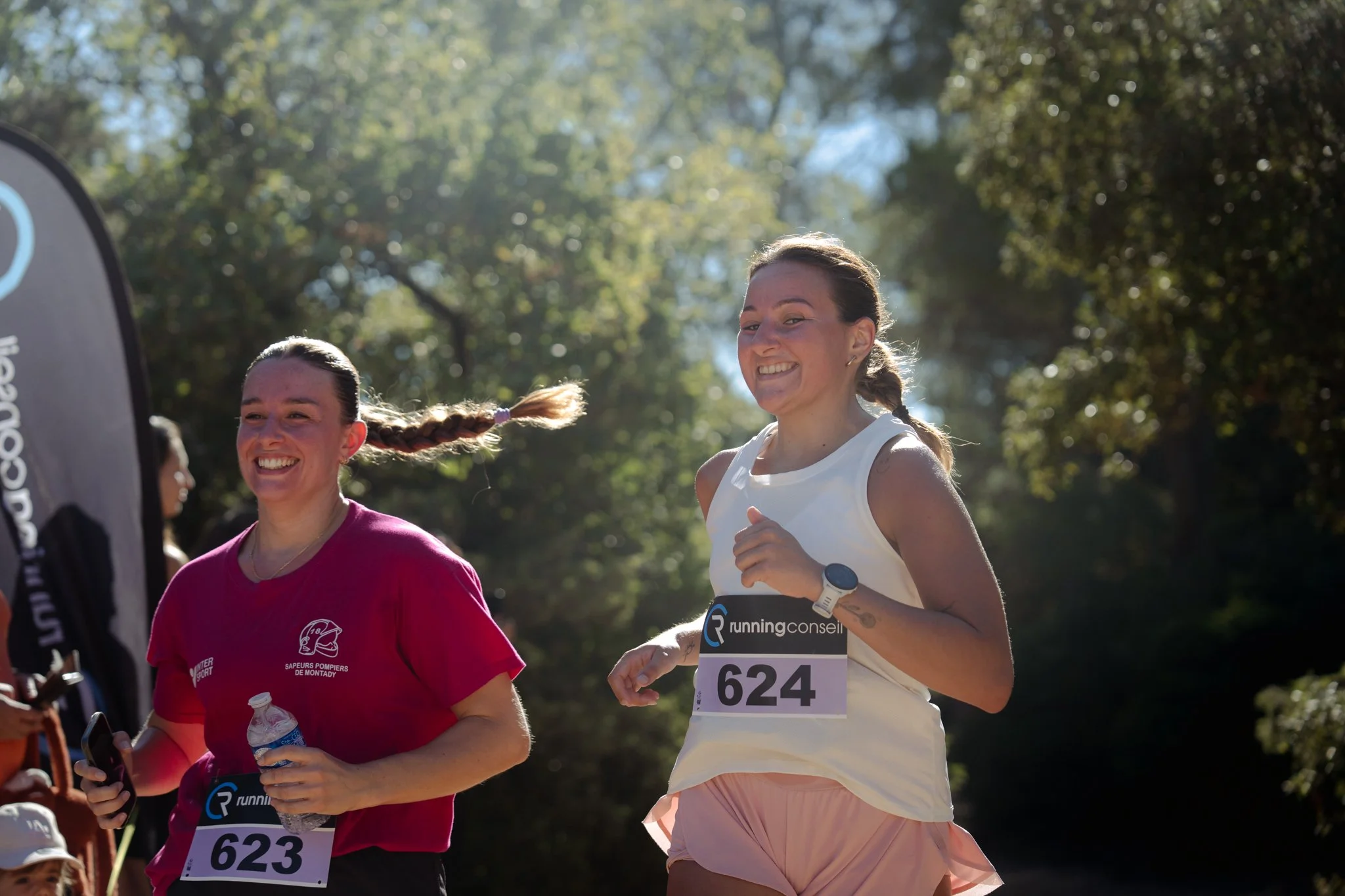 Deux femmes participent à une course de course à pied dans un environnement forestier, souriantes et portant des tenues de sport avec des numéros de course.