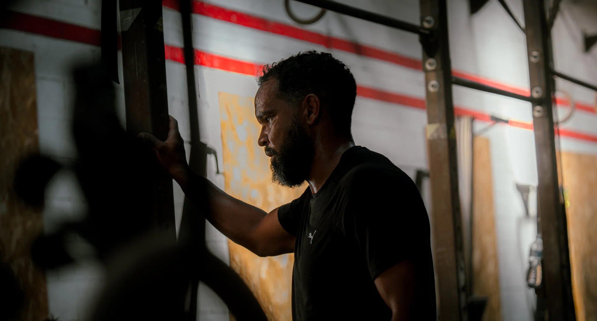 Un homme au visage concentré, les yeux fermés, touchant un mur ou une surface en atelier ou salle de sport, avec des structures métalliques en arrière-plan.
