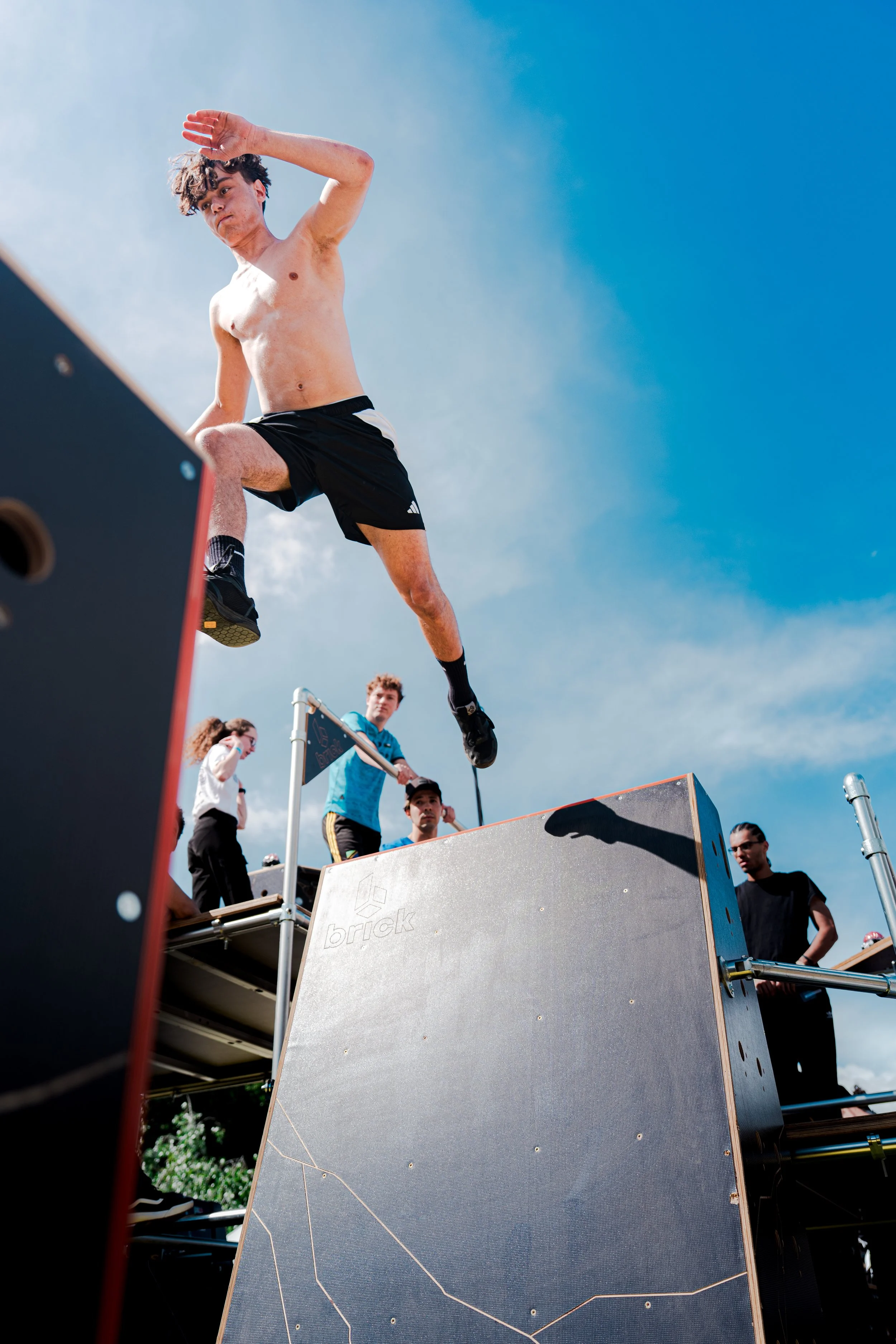 Un jeune homme en short noir sautant lors d'une compétition de parkour en plein air, sous un ciel bleu avec quelques nuages, entouré d'autres personnes qui regardent.
