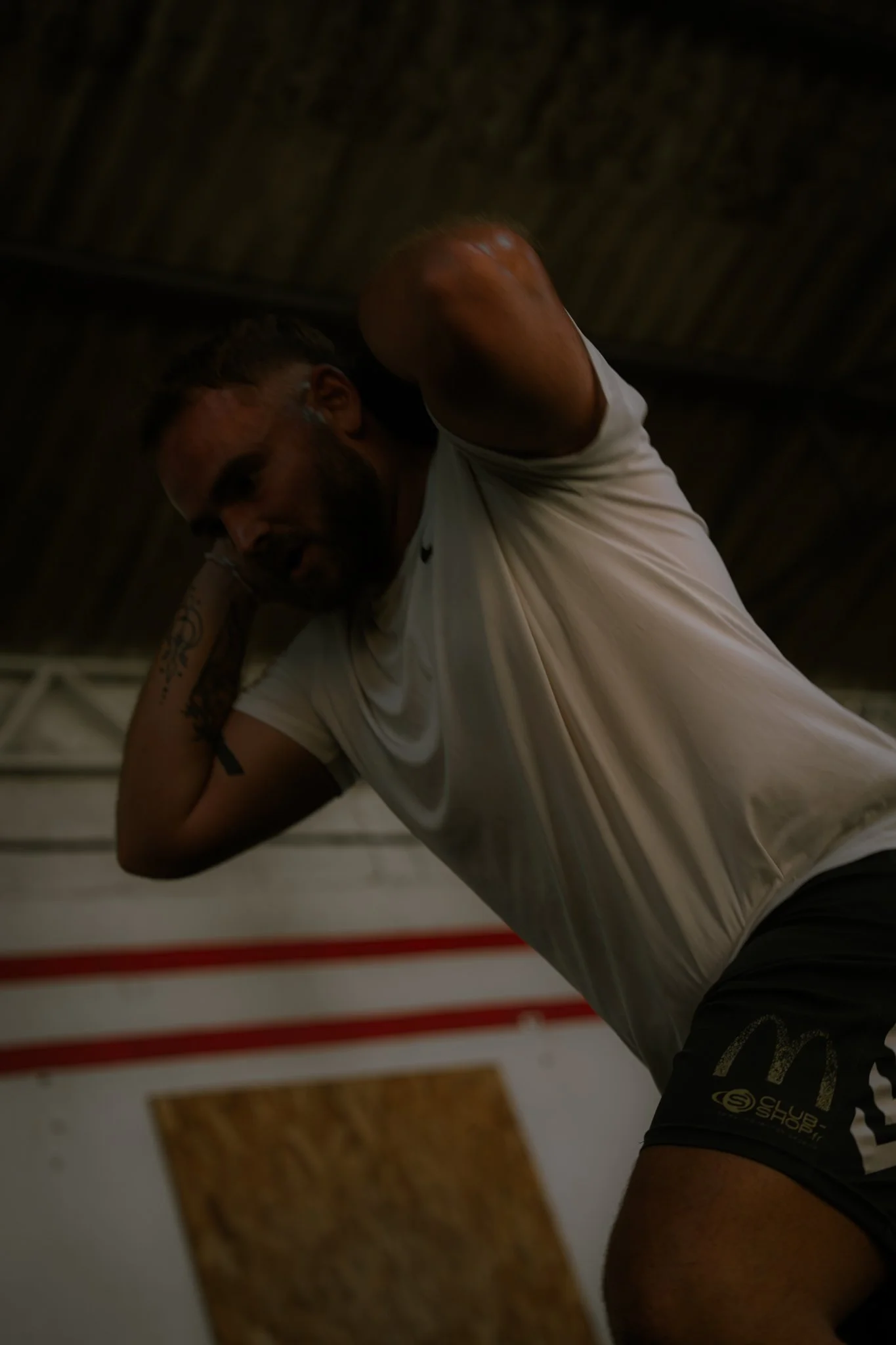 Un homme musclé en maillot de sport blanc s'entraîne dans une salle de sport, avec un plafond en bois visible en arrière-plan.