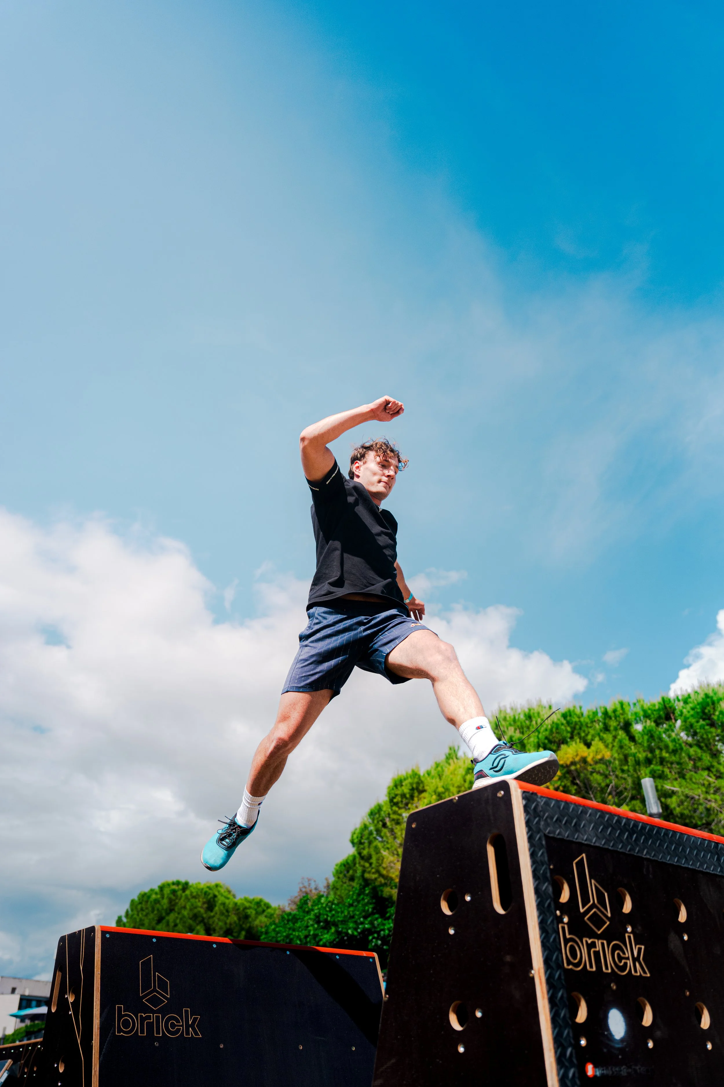 Un jeune homme effectue un saut depuis une plateforme en bois avec le mot 'brick'. Il porte un t-shirt noir, un short bleu et des chaussures de sport, sous un ciel bleu avec des nuages et des arbres verts en arrière-plan.