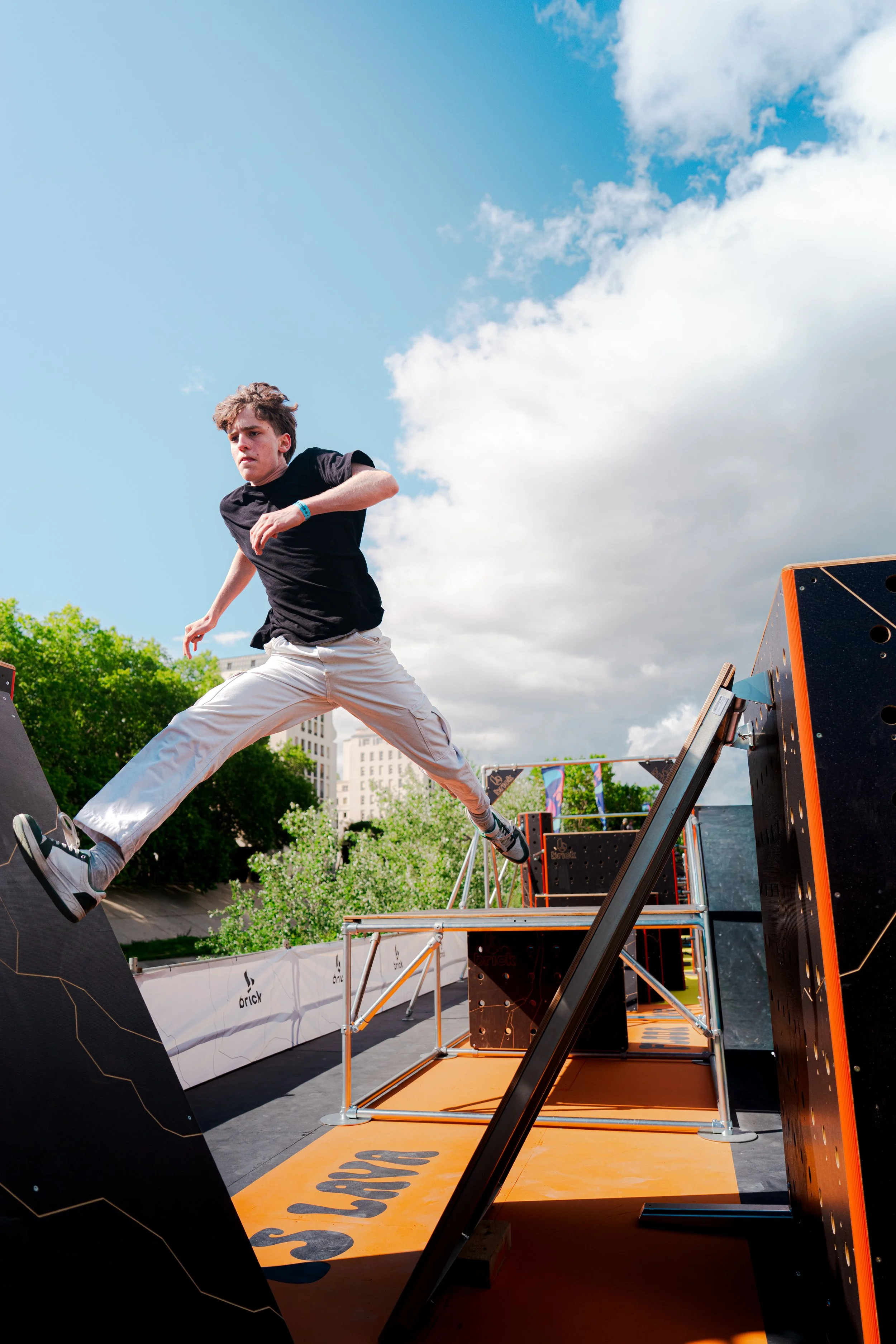 Un jeune homme en t-shirt noir et pantalon clair sautant en l'air à une compétition de sports extrêmes dans un parc urbain, avec un ciel bleu et des bâtiments en arrière-plan.