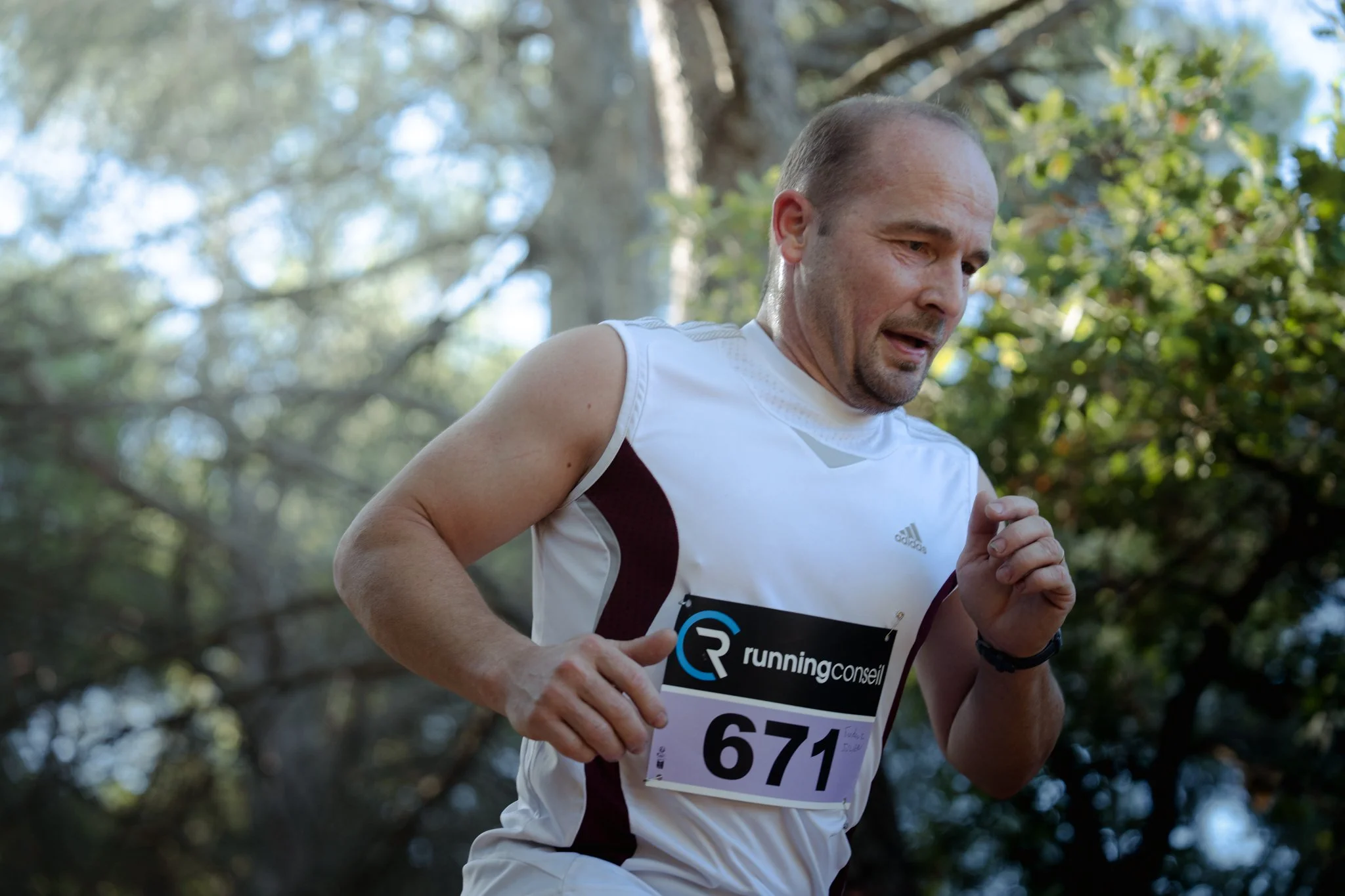 Un homme court dans un parc, portant un numéro de course, en tenue de course blanche. Arbres et ciel en arrière-plan.