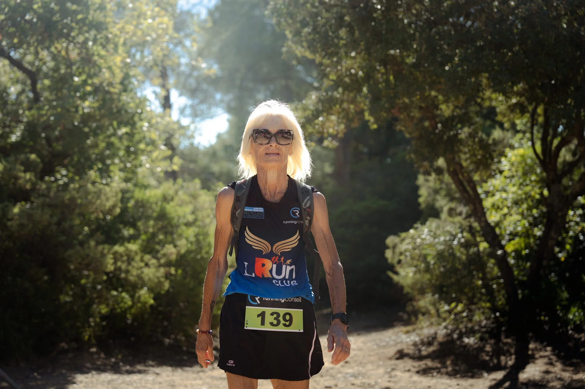 Sénior femme courant dans une forêt en été avec des lunettes de soleil et un numéro de course 139.