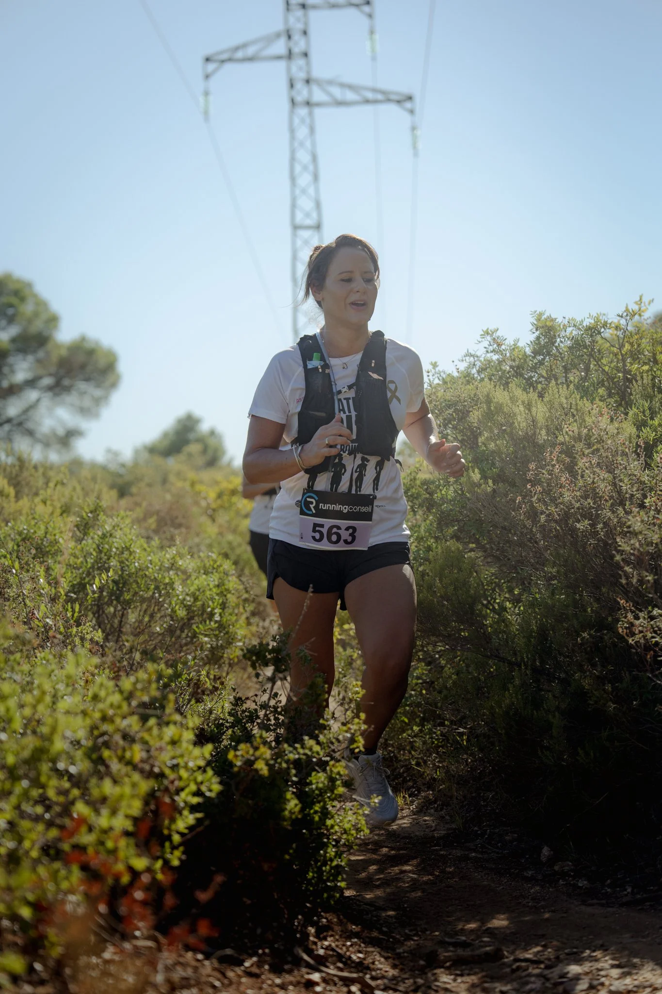 Une femme courant dans un sentier de nature, portant un t-shirt blanc, un sac à dos, et un numéro de course, avec un environnement ensoleillé et des végétations autour.