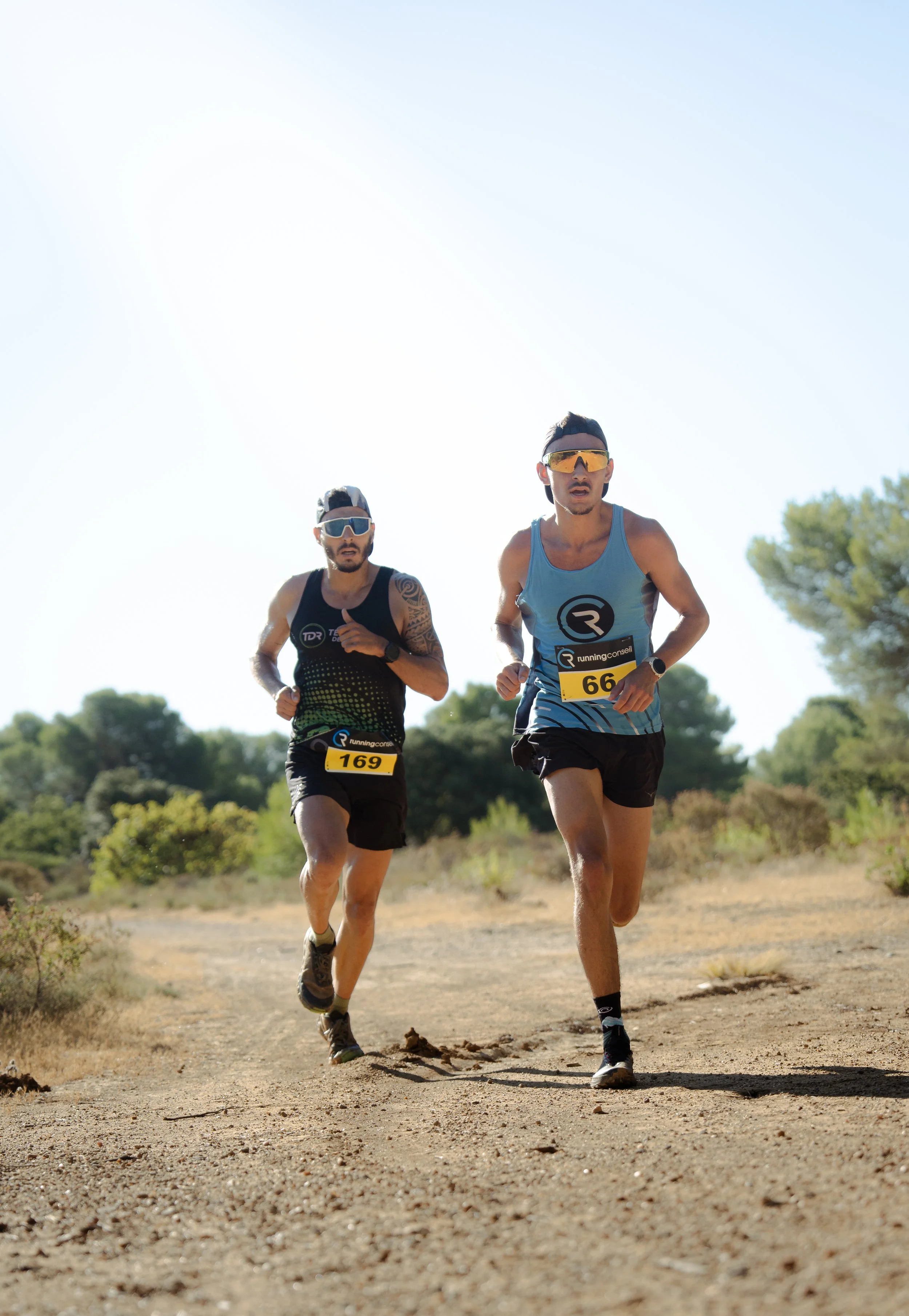 Deux hommes courent lors d'une course en plein air sur un sentier de terre, portant des vêtements de sport et des lunettes de soleil, avec un fond de ciel clair et des arbres en arrière-plan.