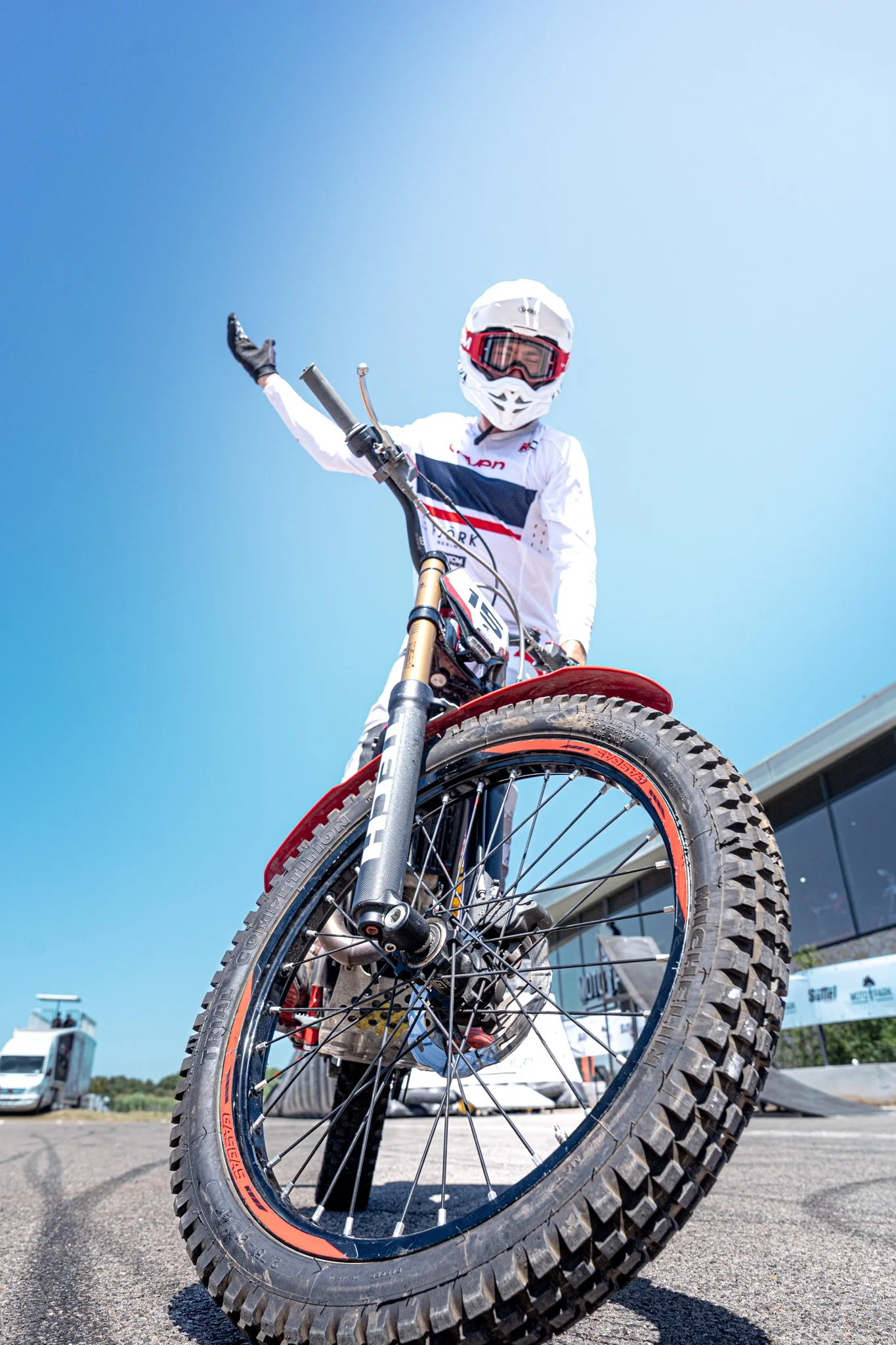 Un cycliste en tenue de compétition, casque et lunettes, sur un vélo de montagne, pris sous un angle très bas avec le pneu en premier plan, dans un espace extérieur en plein jour.