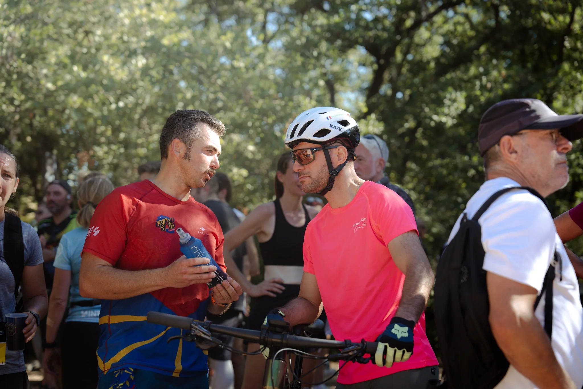 Groupe de personnes lors d'une sortie en plein air, dont deux hommes en conversation, one portant un casque de vélo et des lunettes de soleil, l'autre tenant une bouteille d'eau, avec de nombreux autres individus en arrière-plan.
