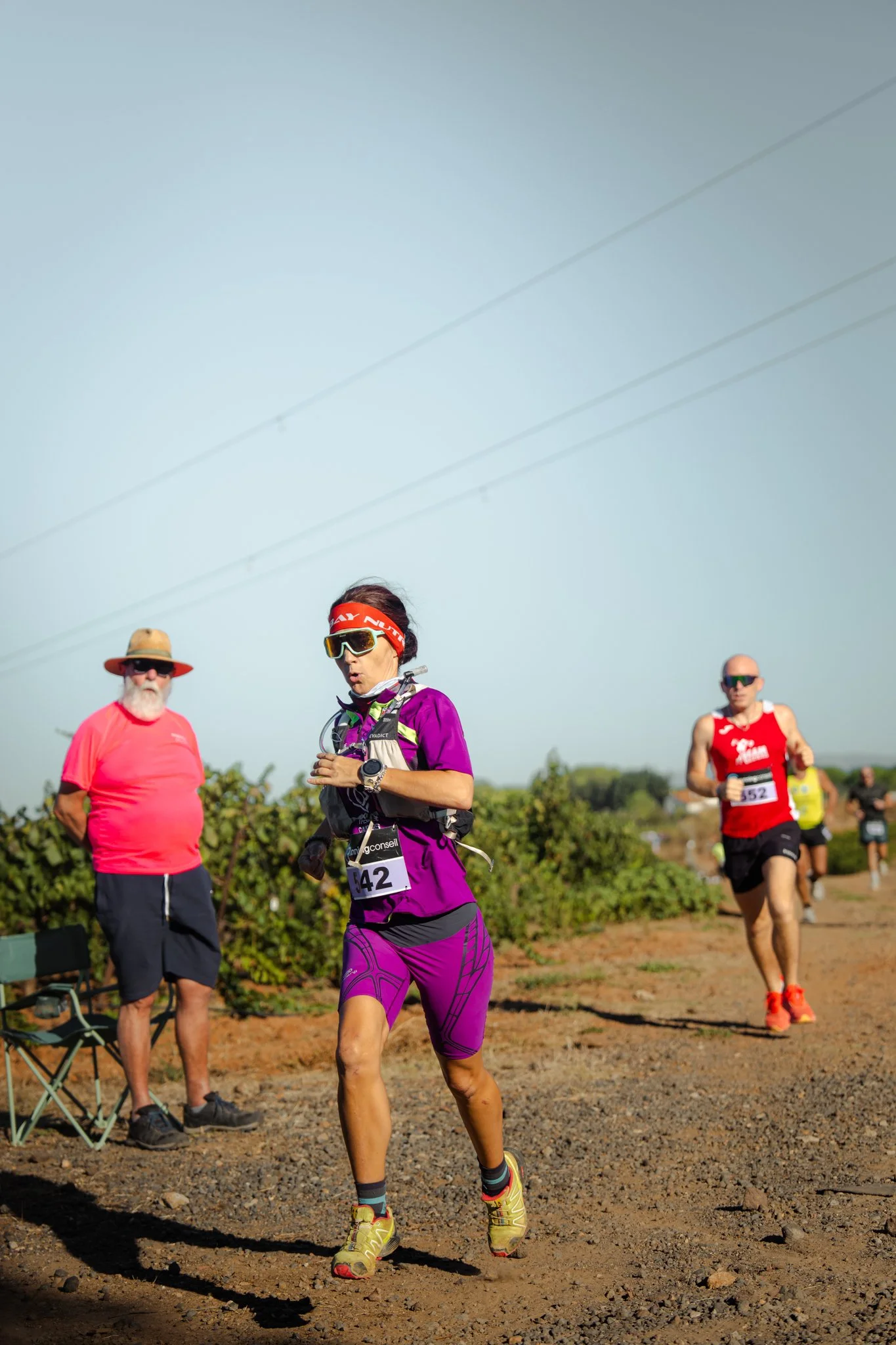 Un groupe de coureurs participate à une course en pleine nature, certains portant des dossards et des vêtements de sport colorés, avec deux personnes âgées debout à côté, observant la course.