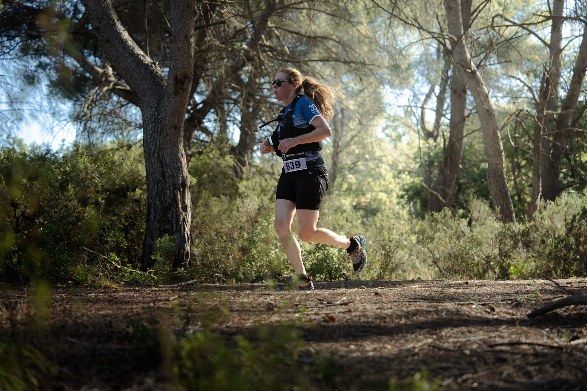 Une femme court dans une forêt, portant un t-shirt bleu, un short noir, des lunettes de soleil et une ceinture d'hydratation, avec un numéro de course 639 attaché à sa taille.