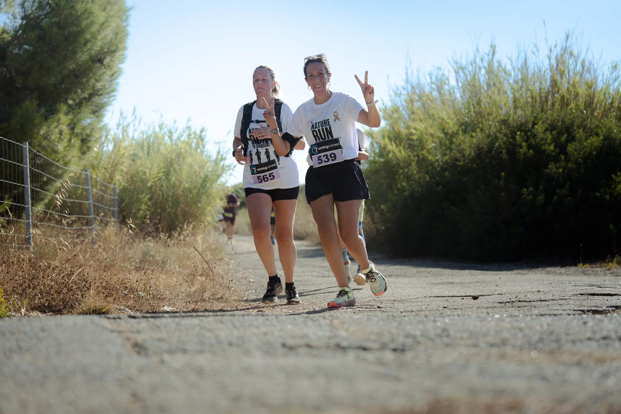 Deux femmes qui participent à une course ou une marche en plein air, portant des t-shirts avec l'inscription "NATURE RUN". Elles portent des numéros de course 565 et 539, et semblent profiter du moment, avec l'une d'elles affichant un signe de paix a