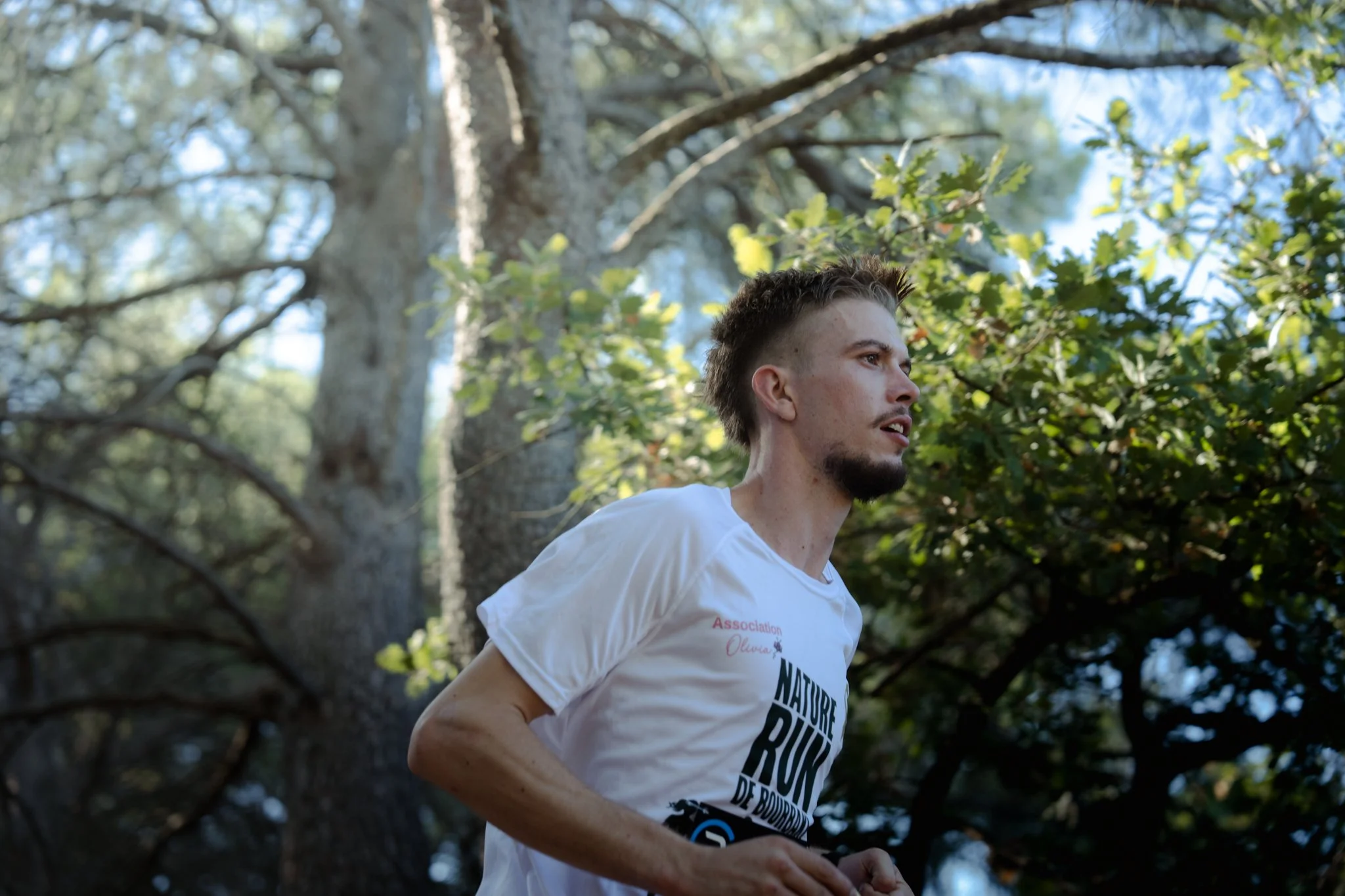 Un homme avec des cheveux courts et une barbe, portant un t-shirt blanc avec du texte, court dans un environnement naturel avec des arbres et des feuilles vertes.