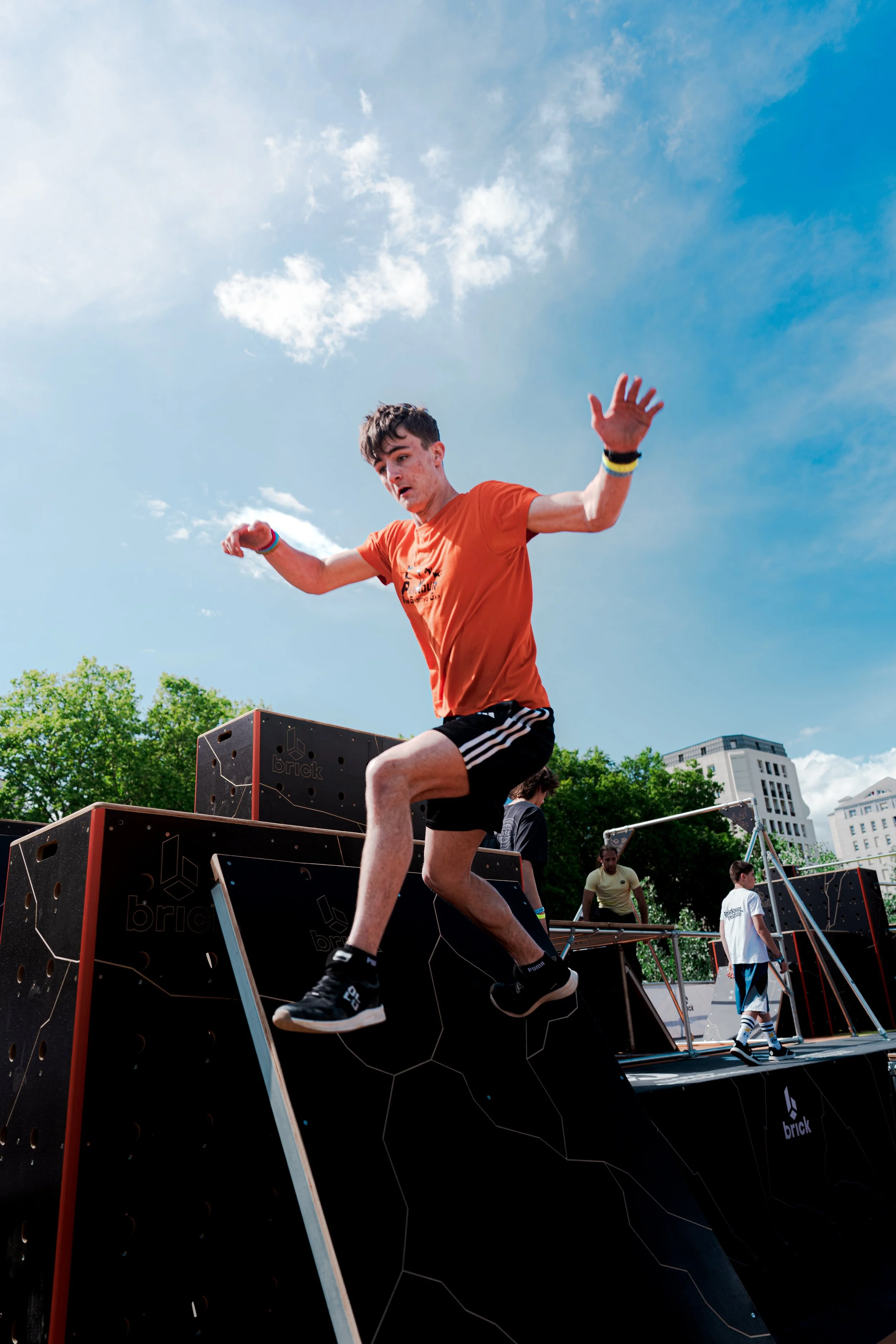 Jeune homme en sprint descente d'une plateforme d'escalade d'obstacles en extérieur, portant un t-shirt orange et des vêtements de sport, avec un ciel bleu en arrière-plan.