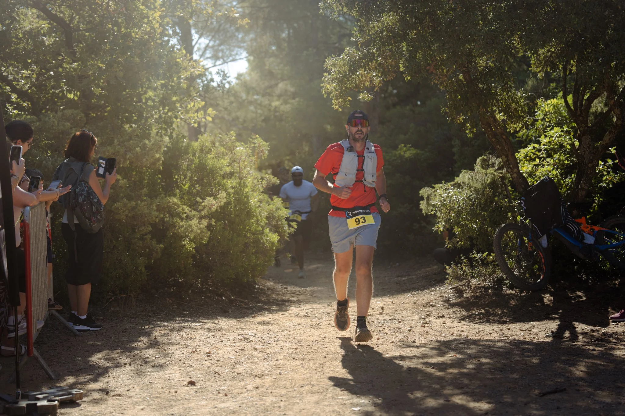 Un homme court dans une course en forêt, portant un t-shirt rouge, un short gris, des lunettes de soleil et un dossard avec le numéro 93. Des spectateurs regardent et prennent des photos sur le côté gauche. Des arbres feuillus entourent le chemin.