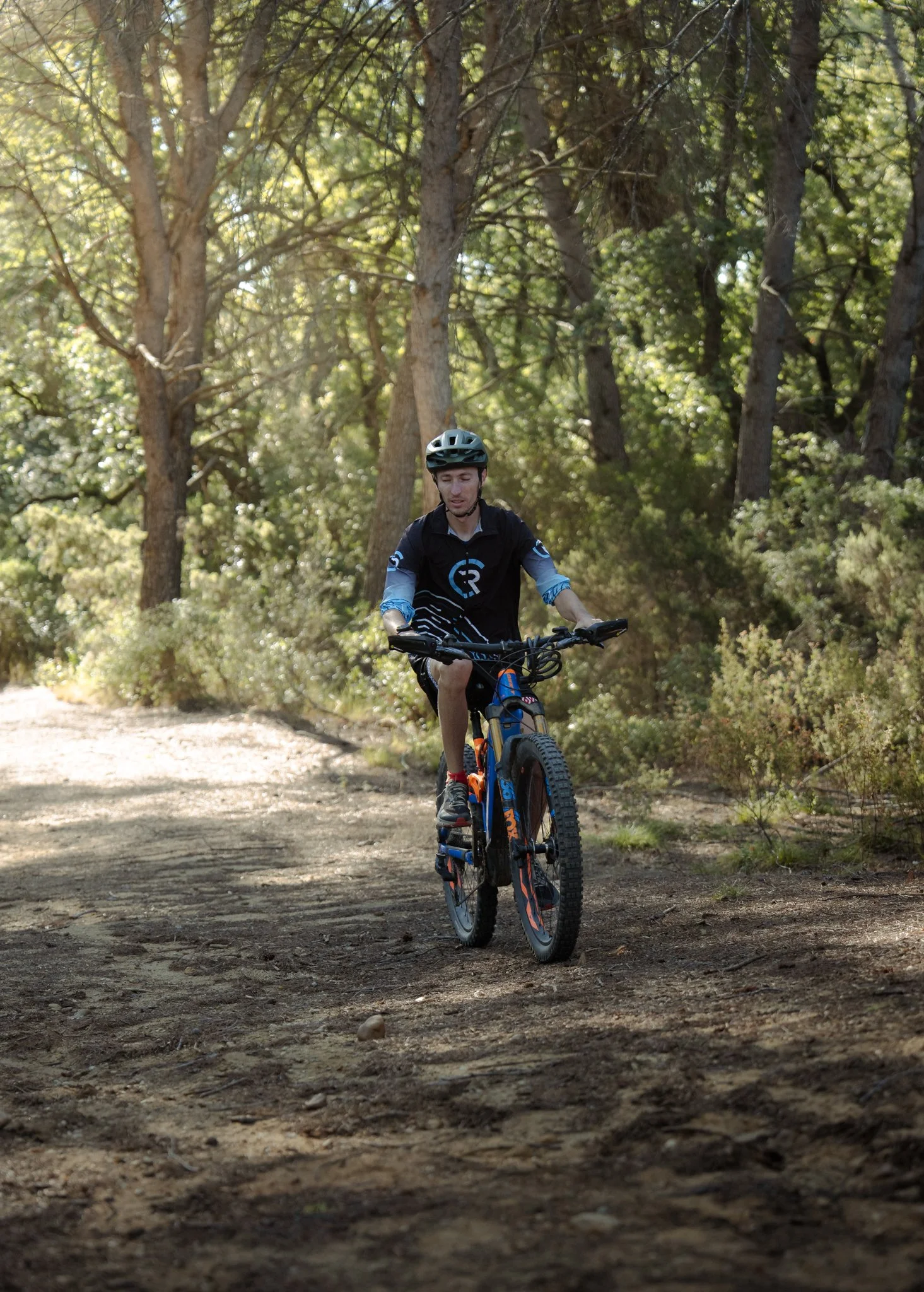 Un homme en tenue de cycliste avec casque, faisant du vélo sur un sentier en forêt, entouré d'arbres et de buissons.