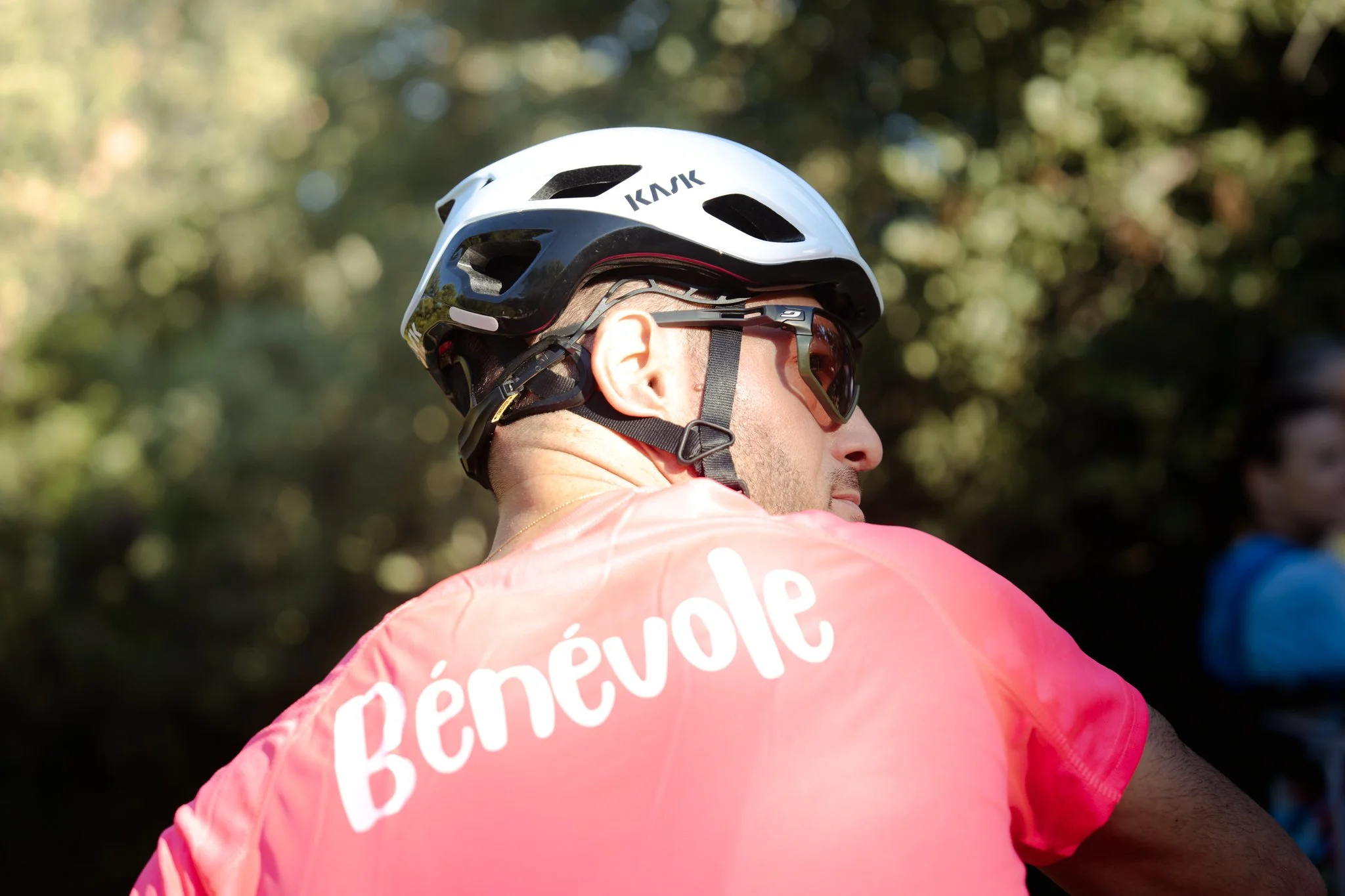 Un homme portant un casque de vélo blanc, des lunettes de protection, et un maillot rose avec du texte blanc.