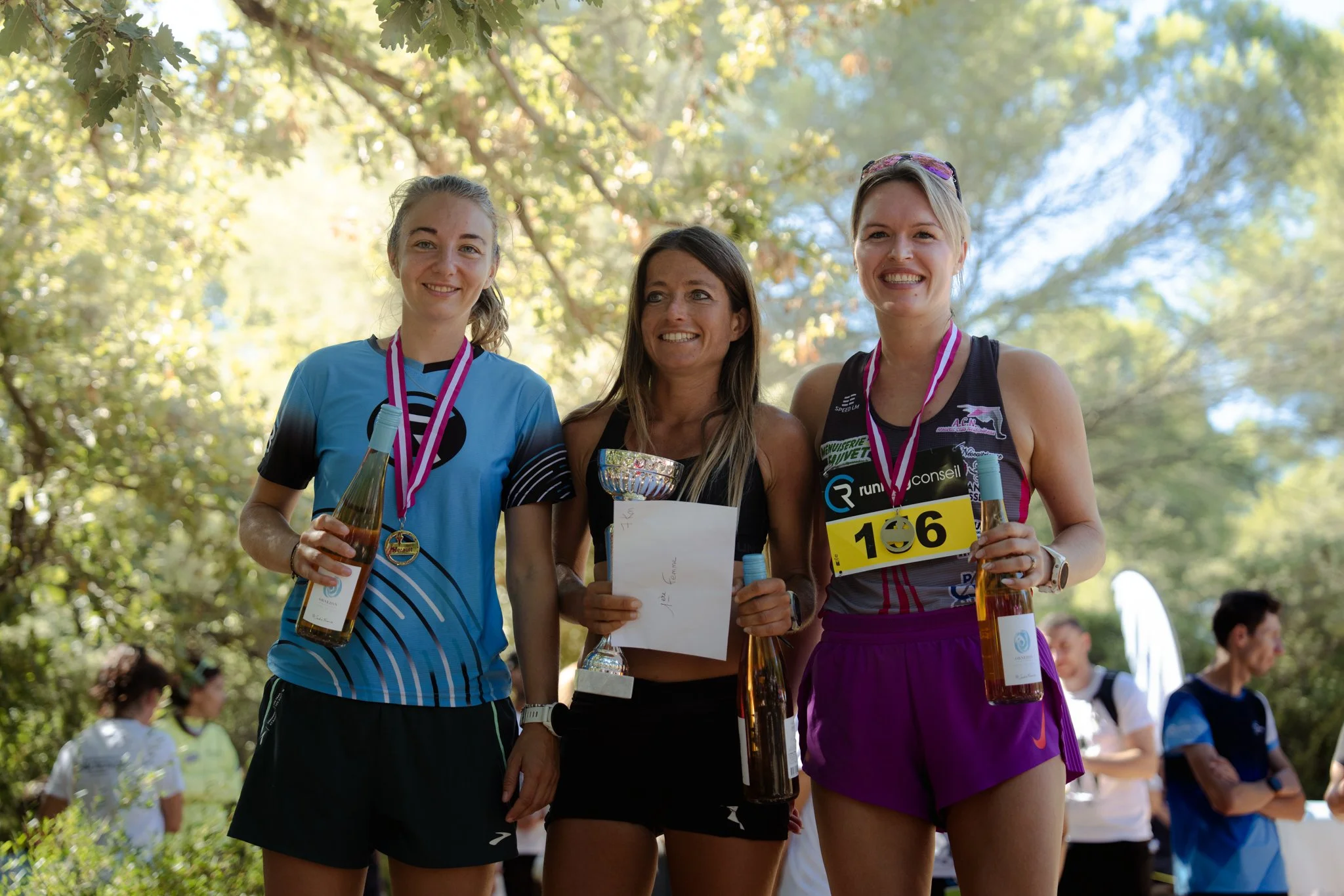 Trois femmes souriantes, portant des médailles, t-shirt de course, détiennent des bouteilles de boisson et un trophée, célèbrent leur victoire lors d'une course en plein air sous un feuillage d'arbres.