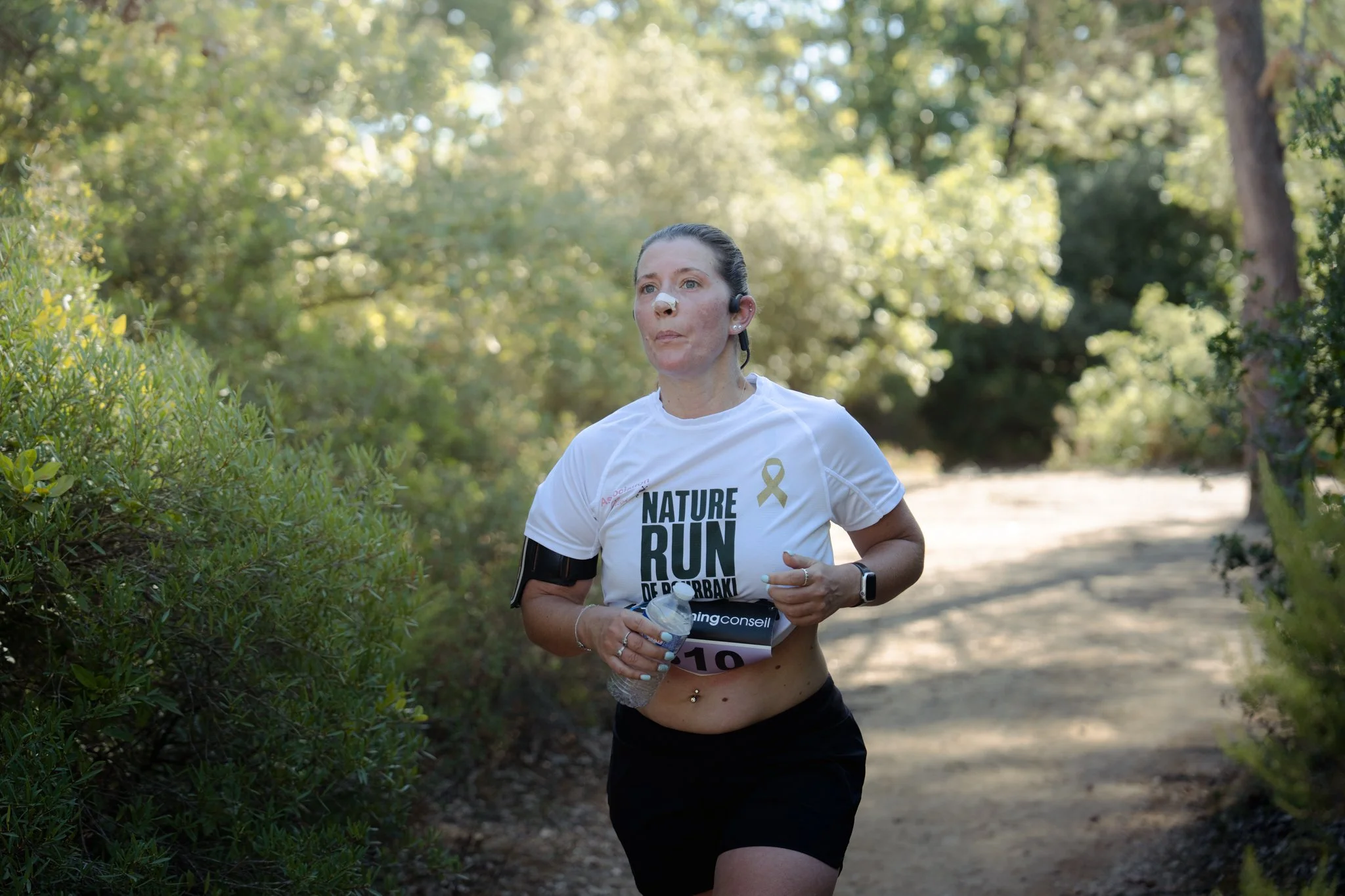 Femme courant dans un parc herbeux avec des arbres, portant un t-shirt blanc avec inscrit 'NATURE RUN' et un bandeau d'oreille Bluetooth.